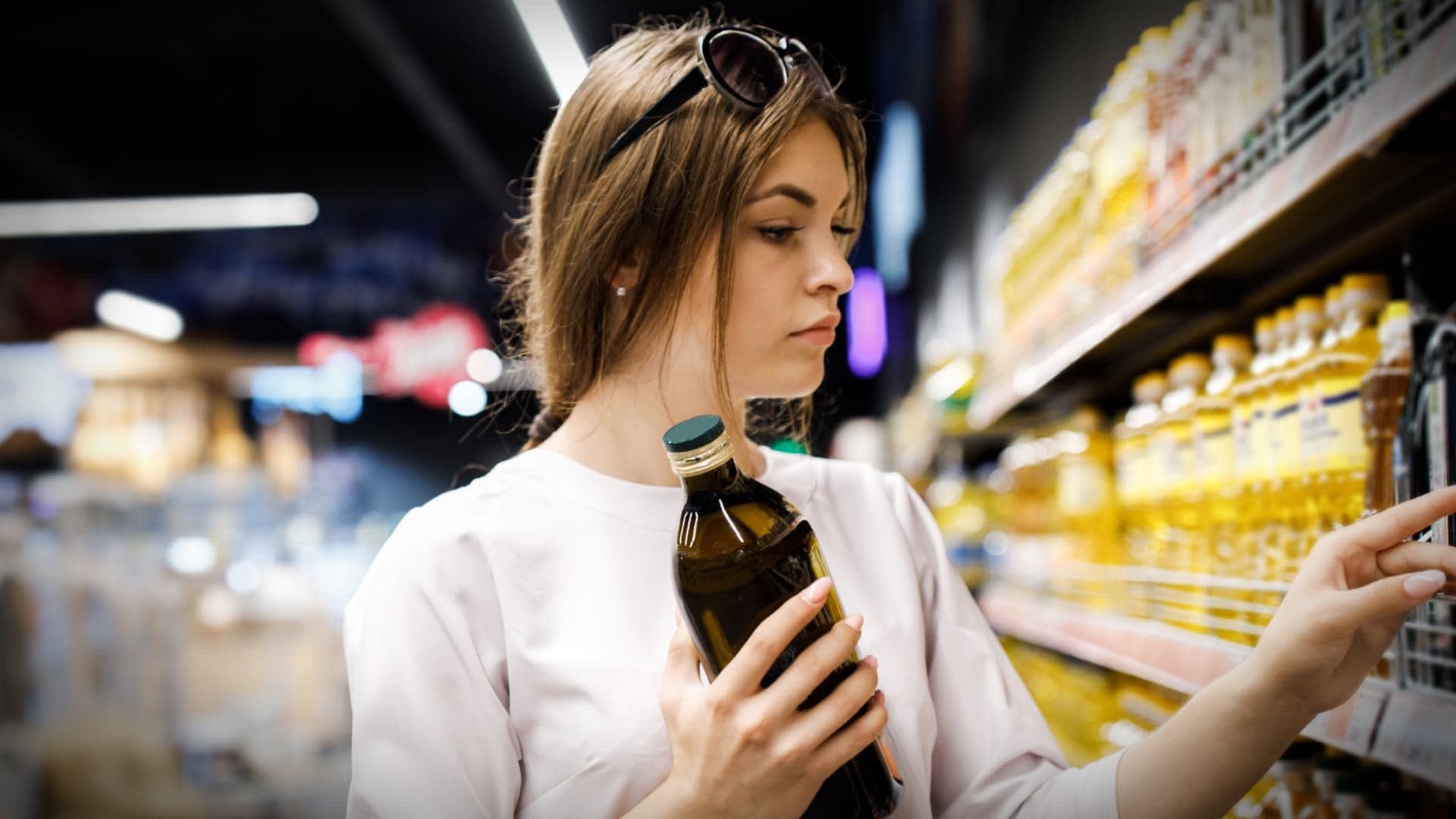 Bir market rafındaki ürünleri incelerken elinde zeytinyağı şişesi tutan kahverengi saçlı kadın. - Olive Oil Times