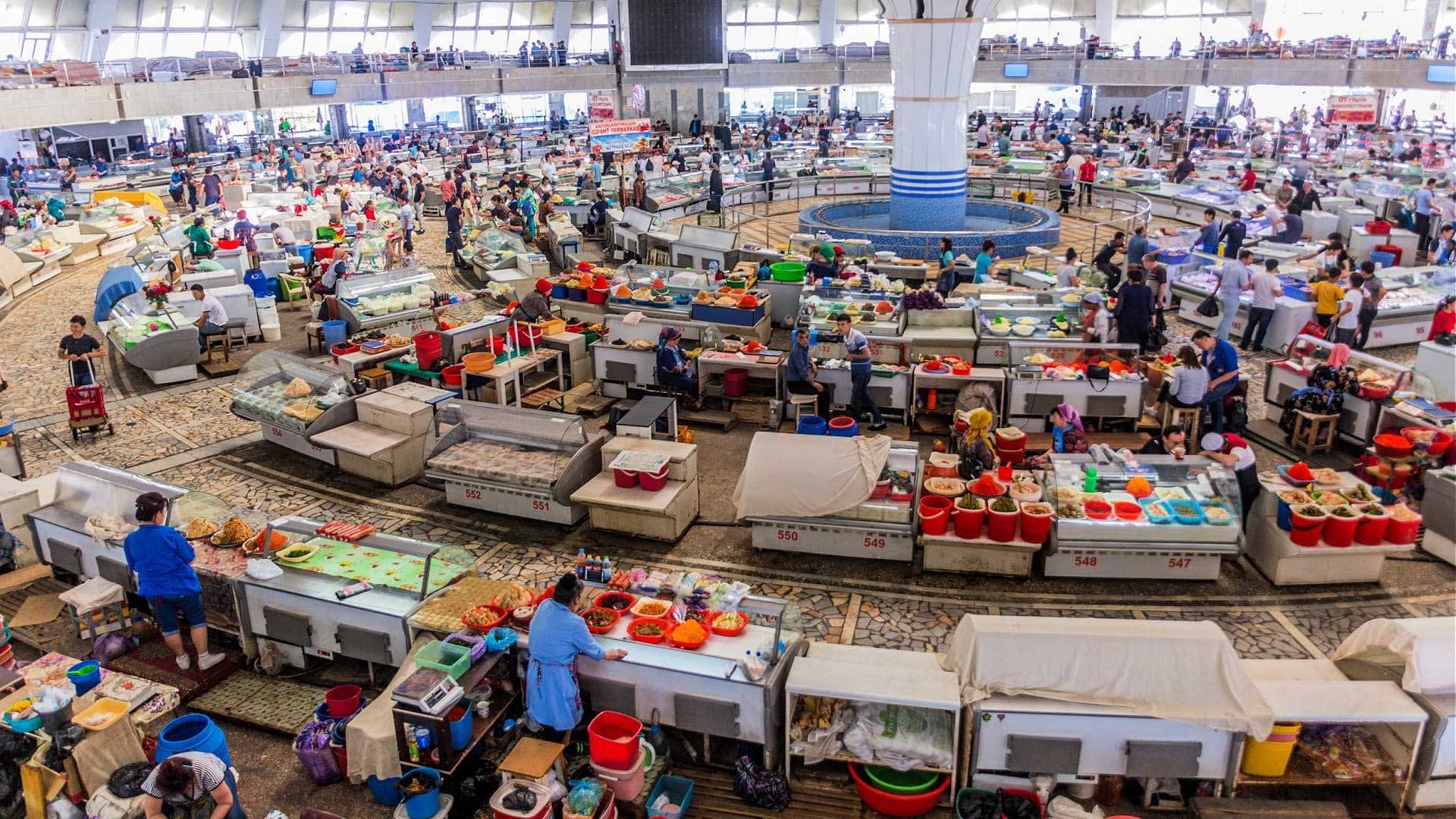 Un mercato vivace, pieno di venditori e clienti, che espone vari stand e prodotti in un ampio spazio al coperto. Olive Oil Times