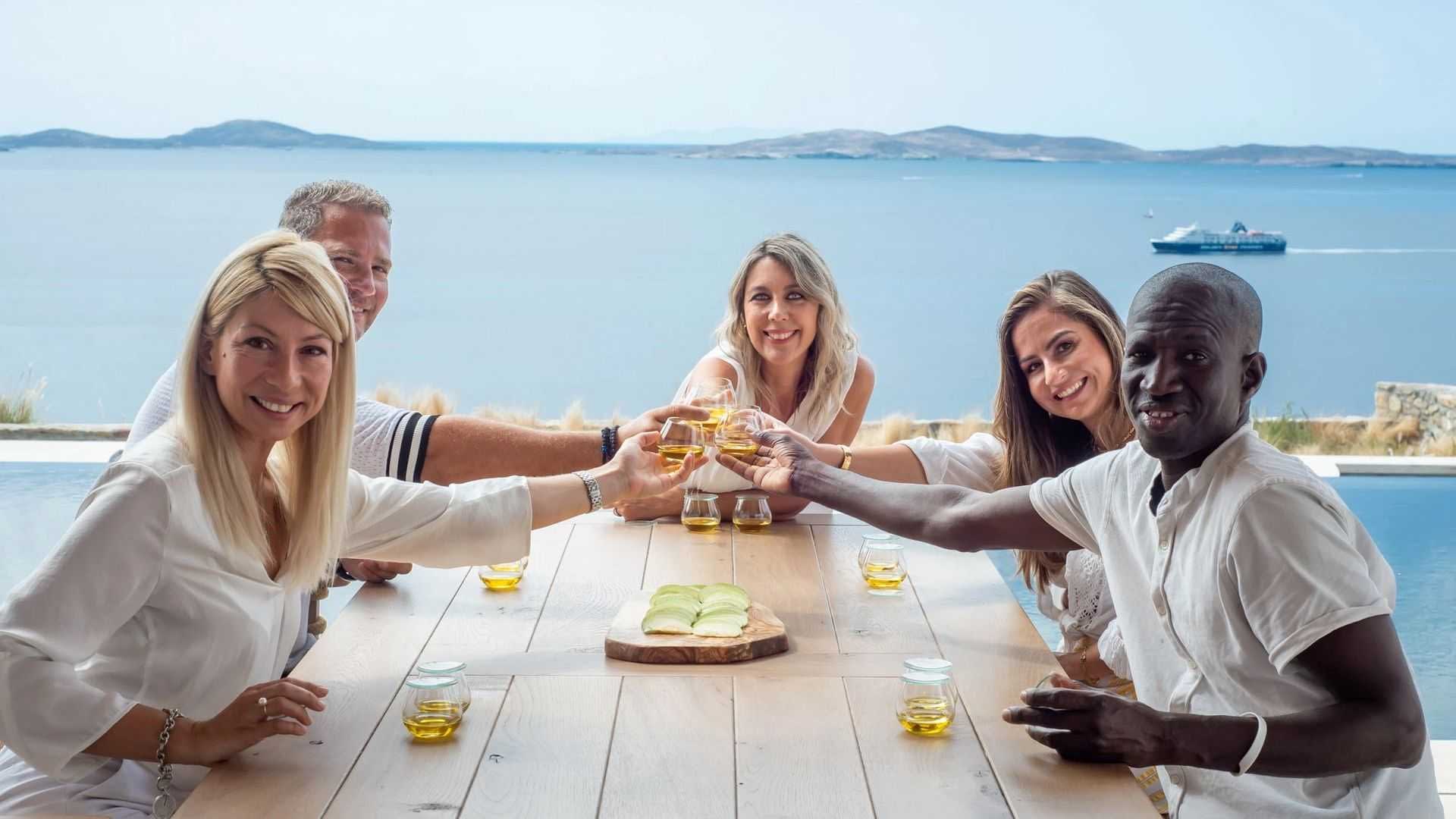 Cinco personas sentadas en una mesa, levantando sus copas para brindar con una vista panorámica del agua de fondo. Olive Oil Times
