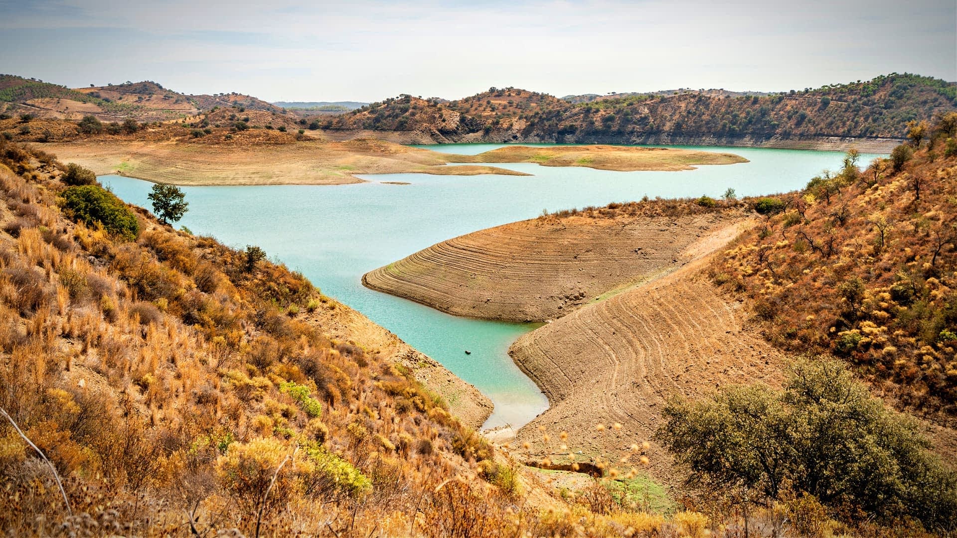Una vista de un embalse con una costa seca y colinas circundantes en un paisaje natural. Olive Oil Times