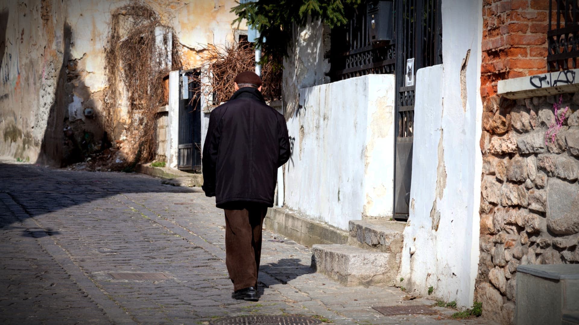 Un anciano camina por una calle adoquinada bordeada de edificios y vegetación. Olive Oil Times