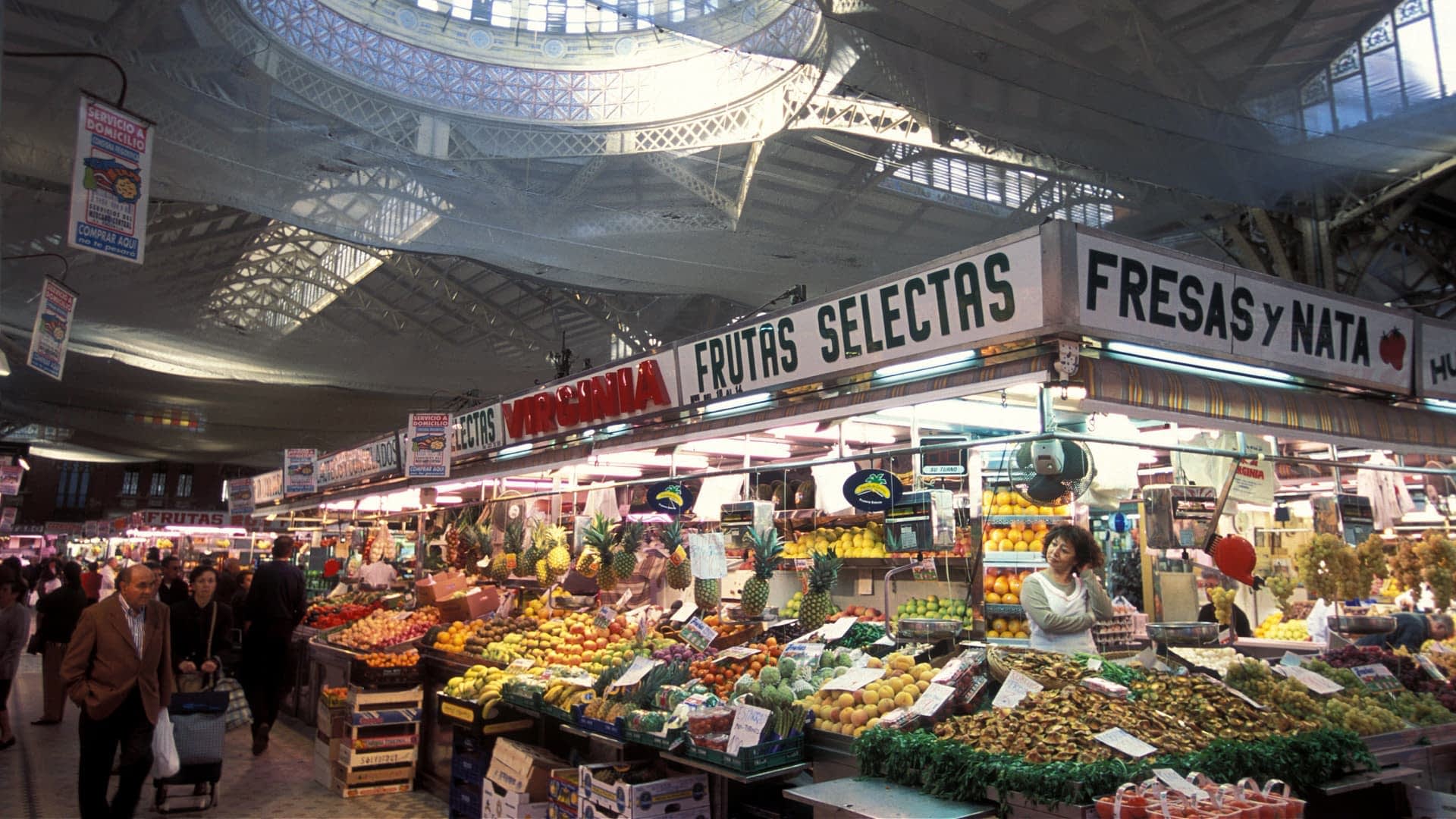 Banca de mercado exibindo uma variedade de frutas e vegetais sob uma grande estrutura de teto. - Olive Oil Times
