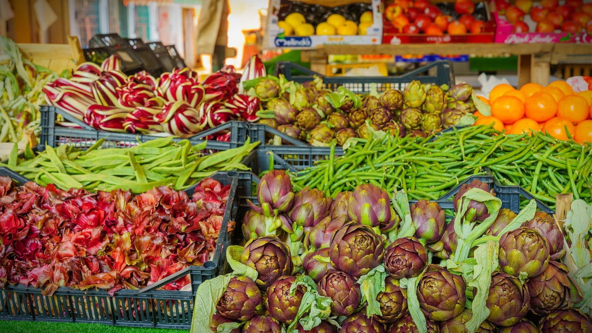Auslage mit verschiedenen frischen Gemüse- und Obstsorten, darunter Artischocken, grüne Bohnen und Orangen auf einem Markt. – Olive Oil Times
