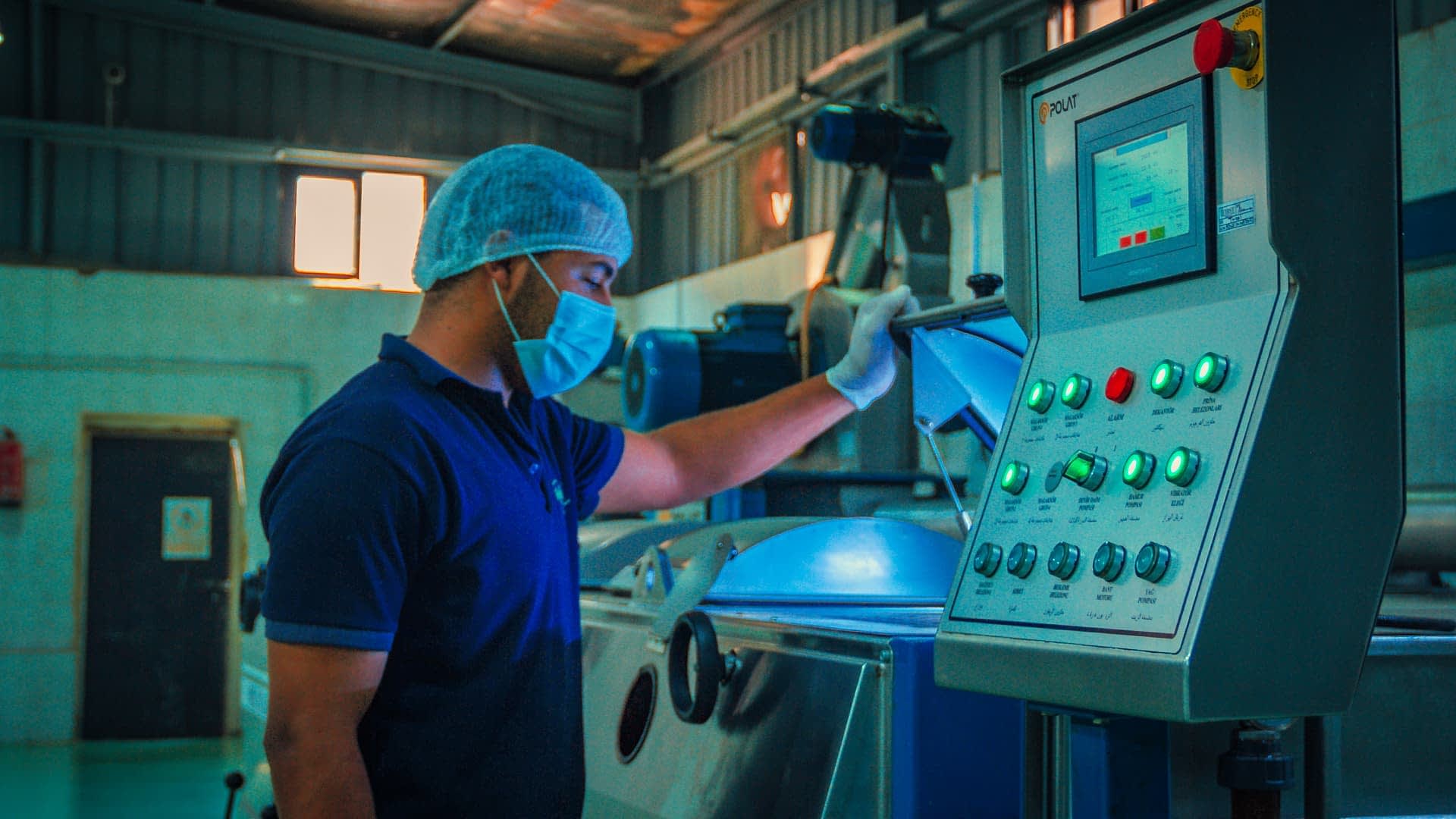 Delavec v modri srajci in mrežici za lase upravlja nadzorno ploščo v industrijskem okolju. - Olive Oil Times