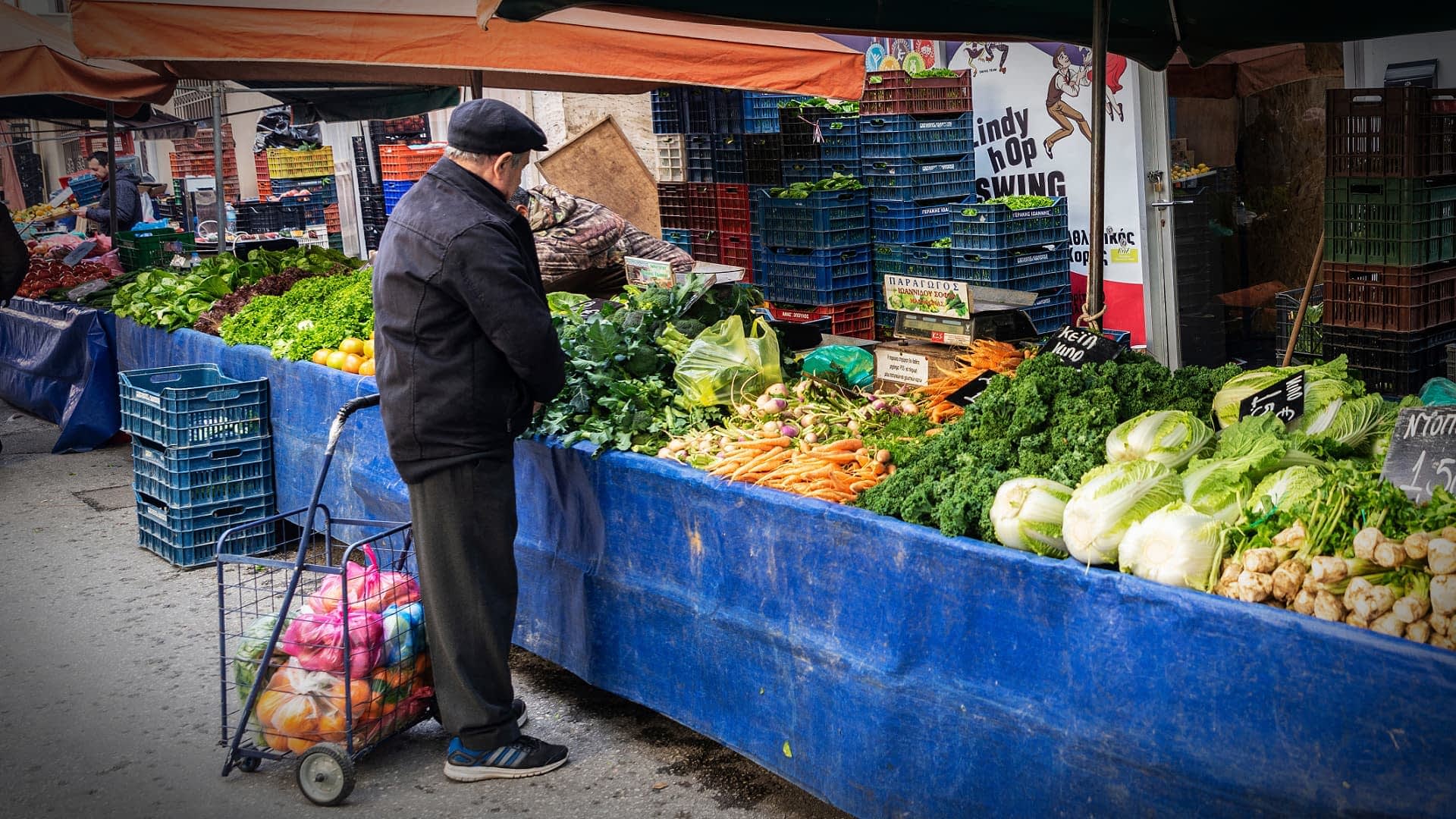 Çeşitli ürünlerin sergilendiği bir pazar tezgahında taze sebzeleri inceleyen adam. - Olive Oil Times