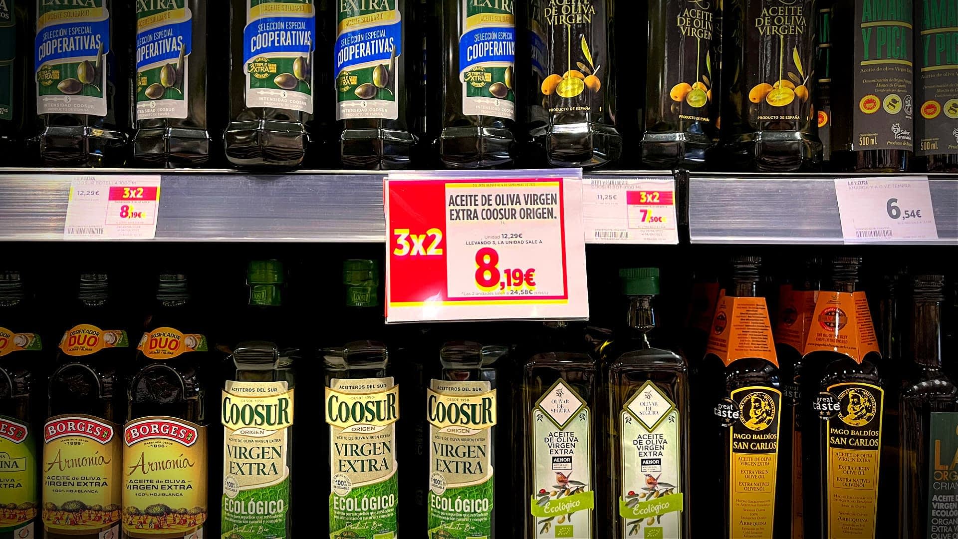 Verschiedene Flaschen Olivenöl stehen mit Preisschildern auf einem Supermarktregal. - Olive Oil Times