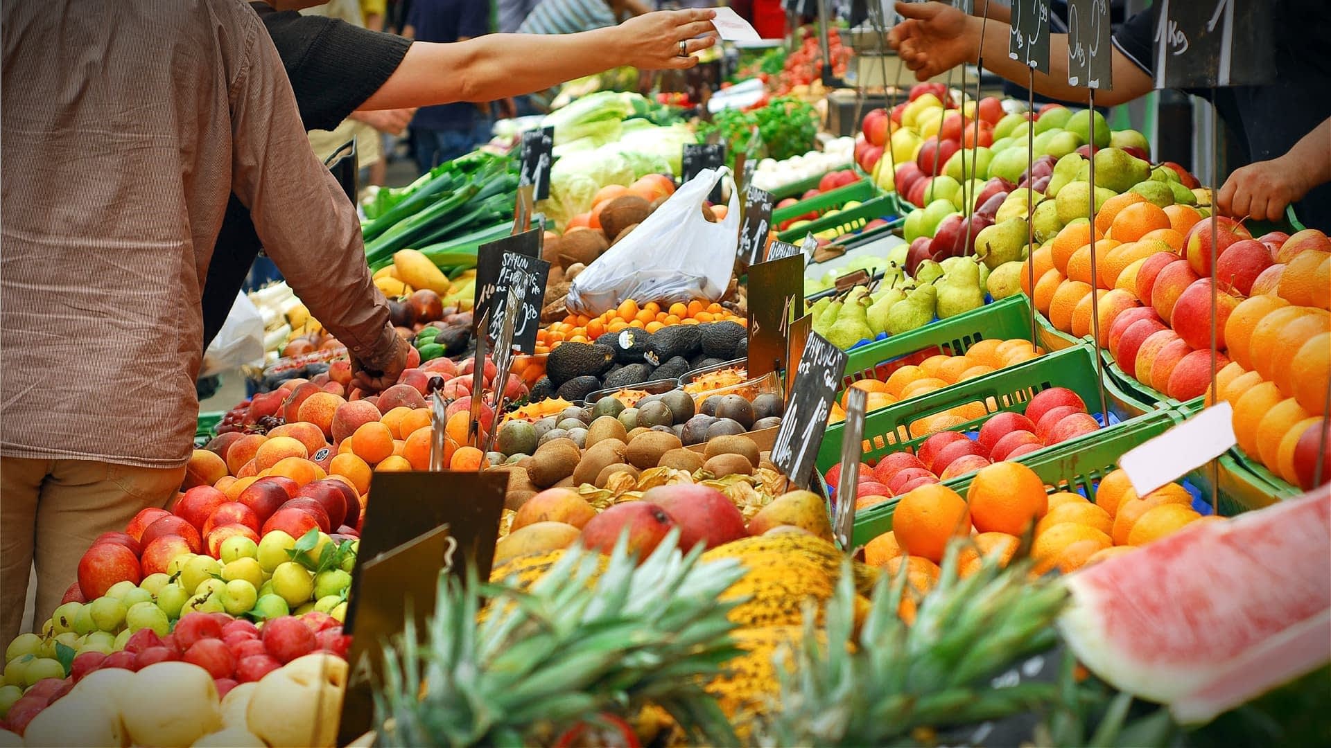 Un étal de marché présentant un large assortiment de fruits frais, notamment des pommes, des oranges et des avocats. Olive Oil Times