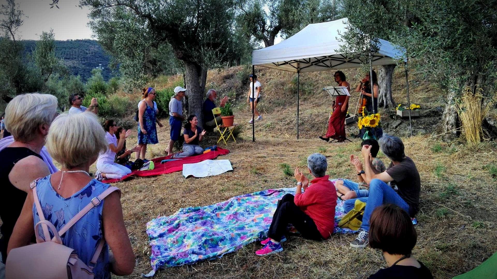 Un gruppo di persone si è radunato all'aperto sotto una tenda per uno spettacolo: alcuni erano seduti su coperte e altri in piedi. Olive Oil Times
