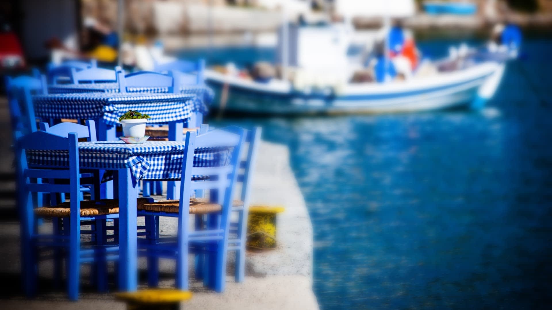 Σετ μπλε ξύλινες καρέκλες και ένα τραπέζι με ένα καρό τραπεζομάντιλο κοντά σε ένα σώμα νερού. - Olive Oil Times