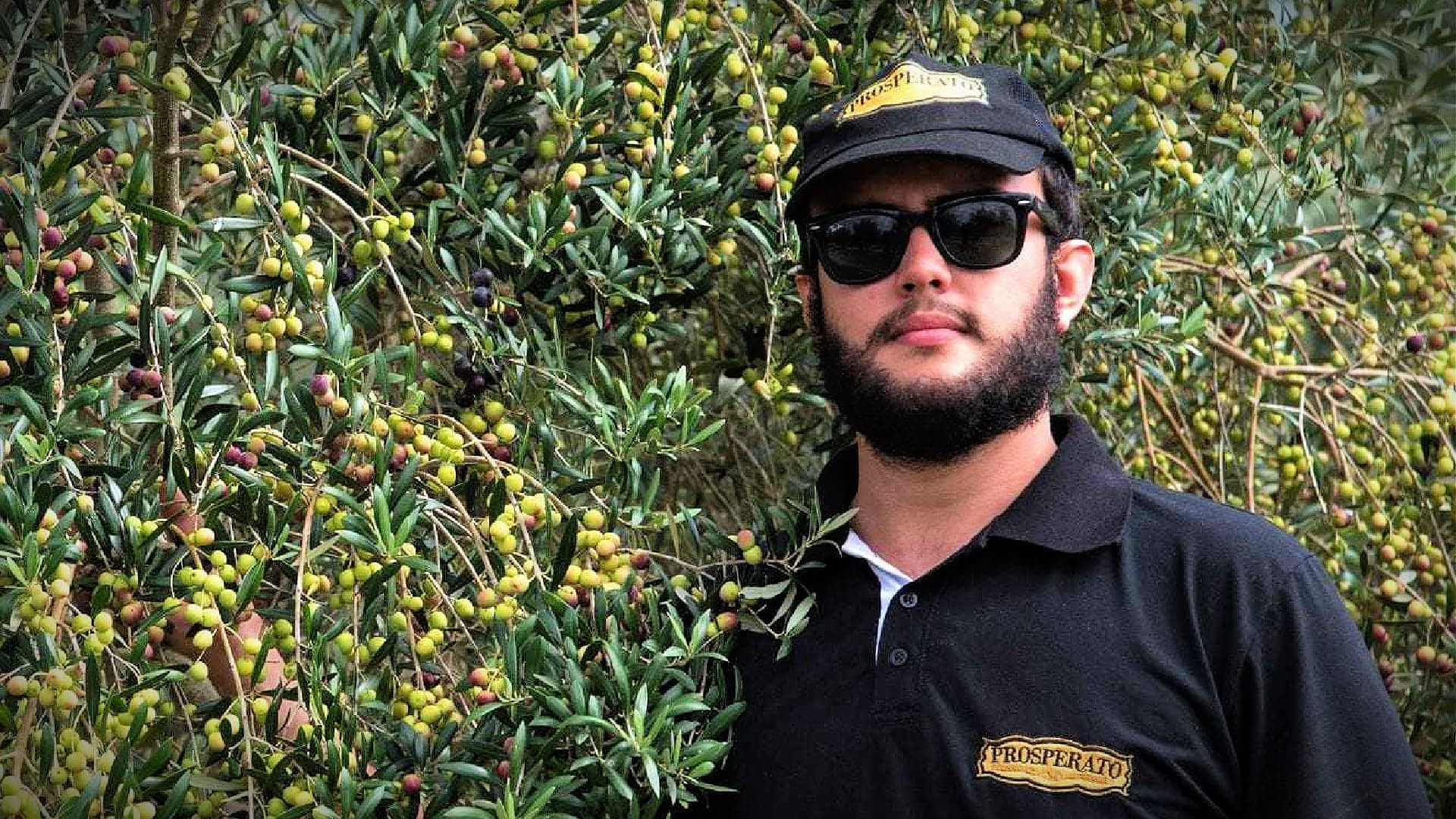 Uomo con occhiali da sole e berretto nero in piedi accanto a un ulivo con olive verdi. - Olive Oil Times