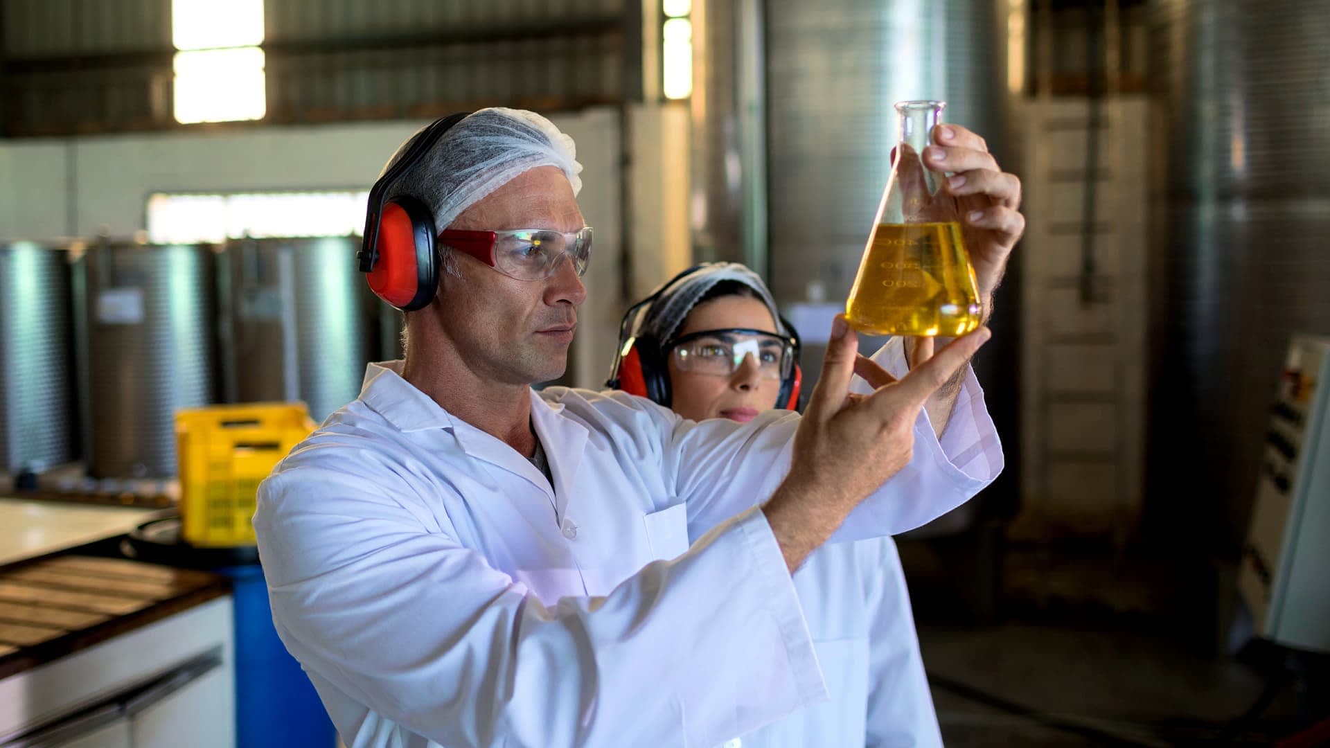 Twee laboratoriummedewerkers in witte jassen onderzoeken een kolf met gele vloeistof in een laboratoriumomgeving. Olive Oil Times