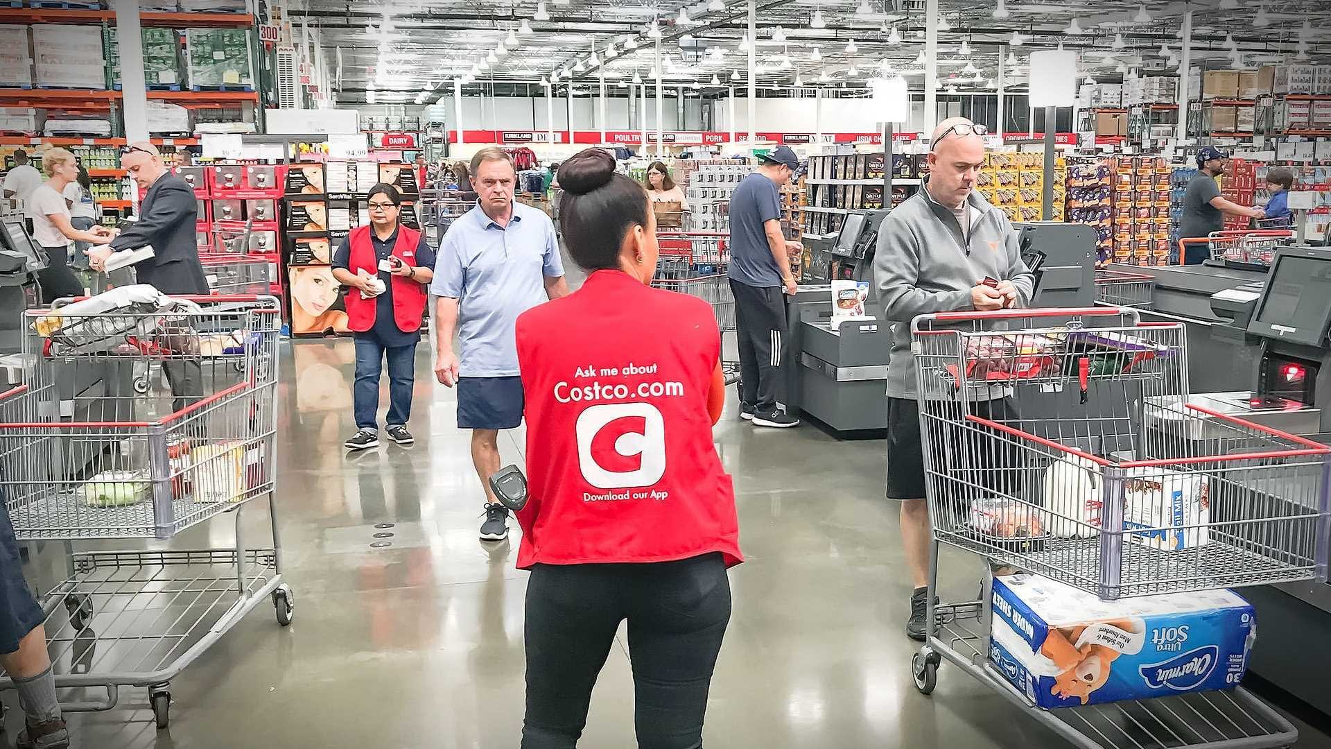 Женщина в красном жилете стоит в кассовой зоне магазина Costco на фоне тележек и покупателей. Olive Oil Times