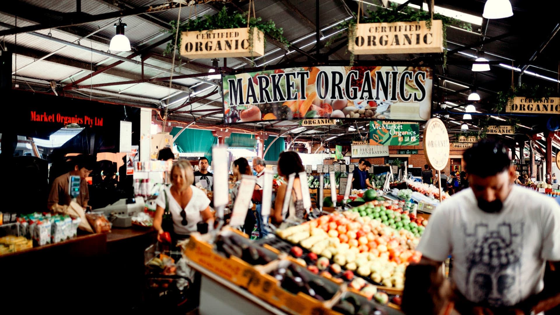 Πινακίδα που γράφει "Market Organics" σε μια πολυσύχναστη αγορά αγροτών με διάφορα βιολογικά προϊόντα που εκτίθενται. - Olive Oil Times