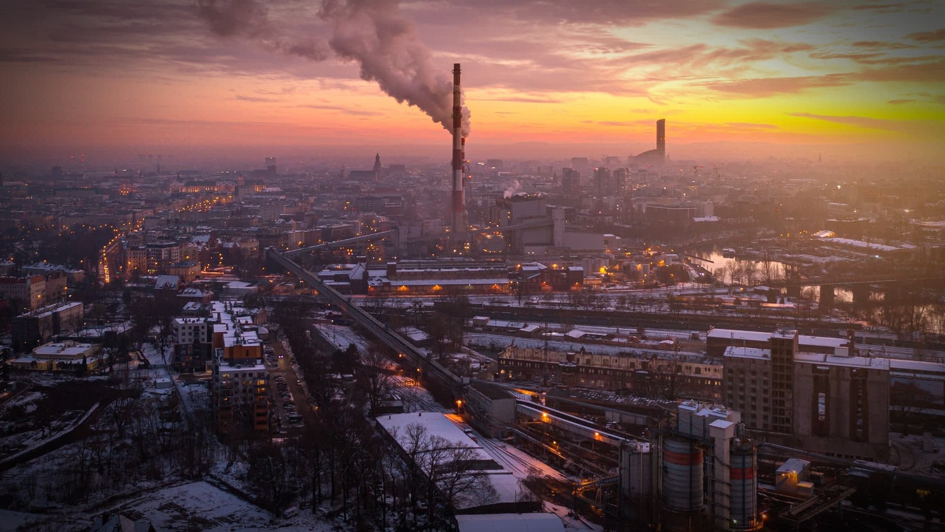 Luchtfoto van een industriegebied met schoorstenen die rook uitstoten tegen de ondergaande zon. Olive Oil Times