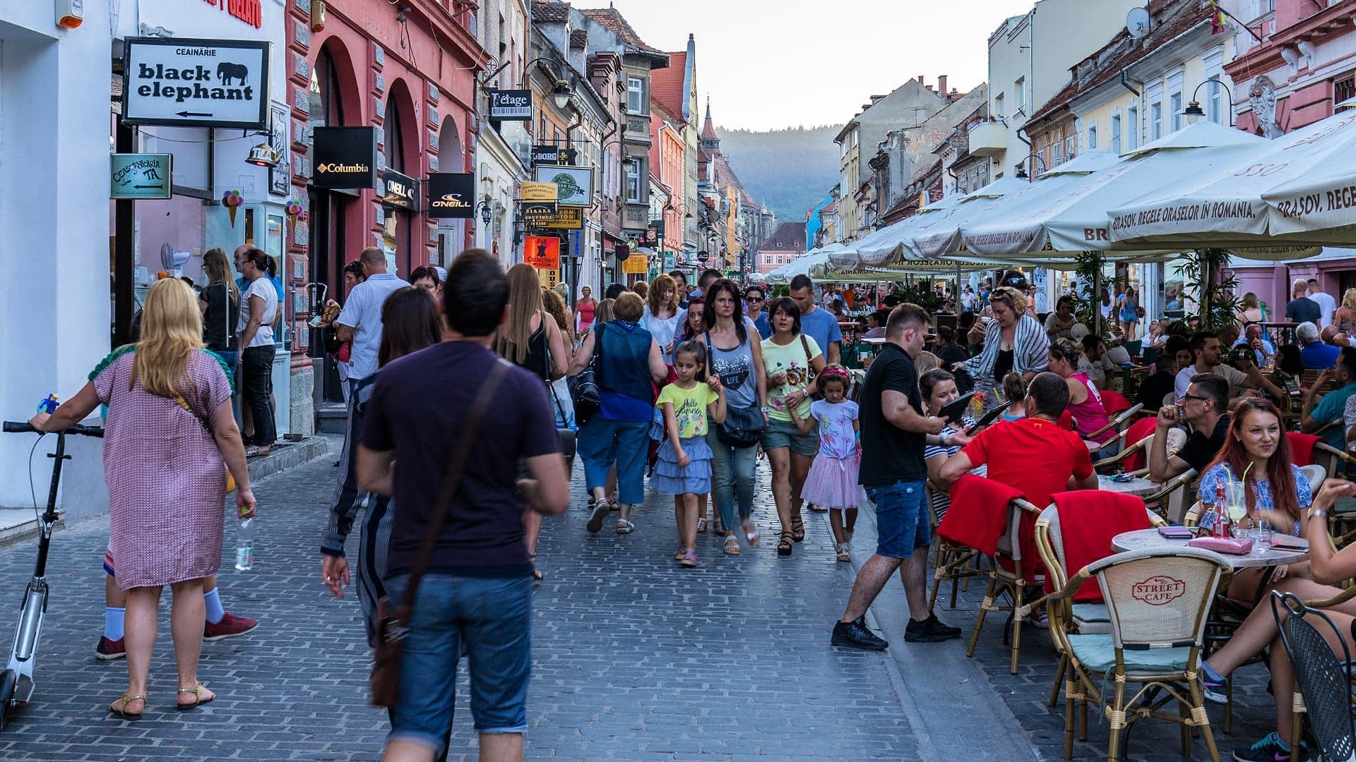 Množica ljudi, ki hodi po ulici z zunanjimi sedeži in trgovinami v mestu. - Olive Oil Times