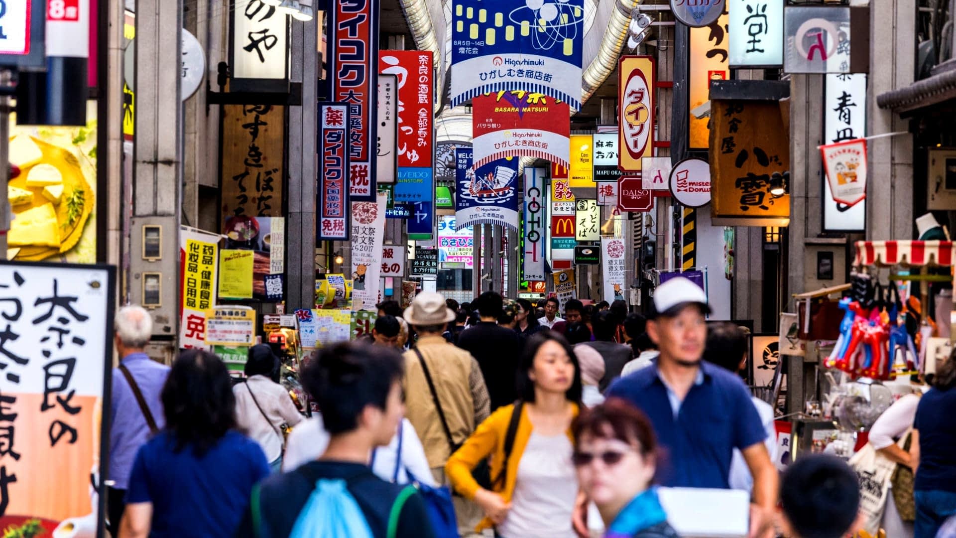 Japonya'da rengarenk tabelalarla dolu hareketli bir sokak pazarında yürüyen insan kalabalığı. - Olive Oil Times