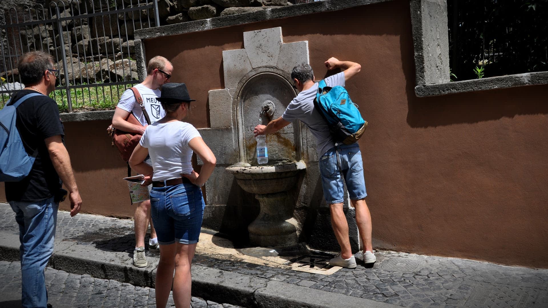 Groupe de personnes interagissant avec une fontaine murale dans un cadre extérieur. - Olive Oil Times