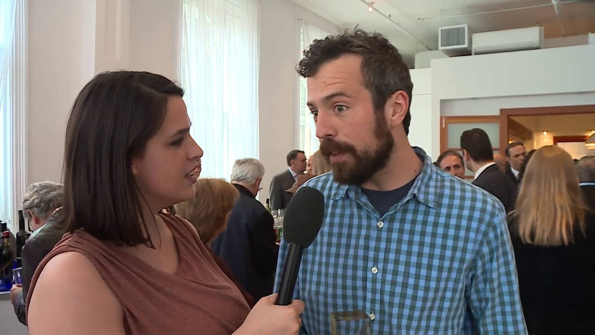 Ein Mann in einem blau karierten Hemd spricht bei einer Veranstaltung mit einer Frau, die ein Mikrofon hält. - Olive Oil Times