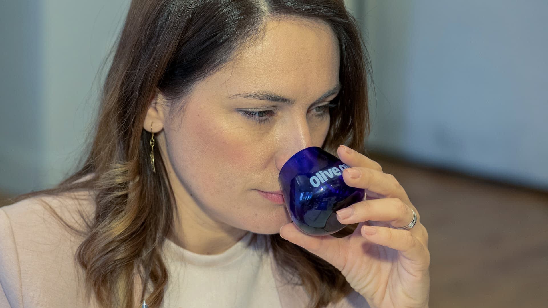 Frau mit braunem Haar hält ein blaues Verkostungsglas mit der Aufschrift „Olivenöl“ nah an ihr Gesicht. – Olive Oil Times