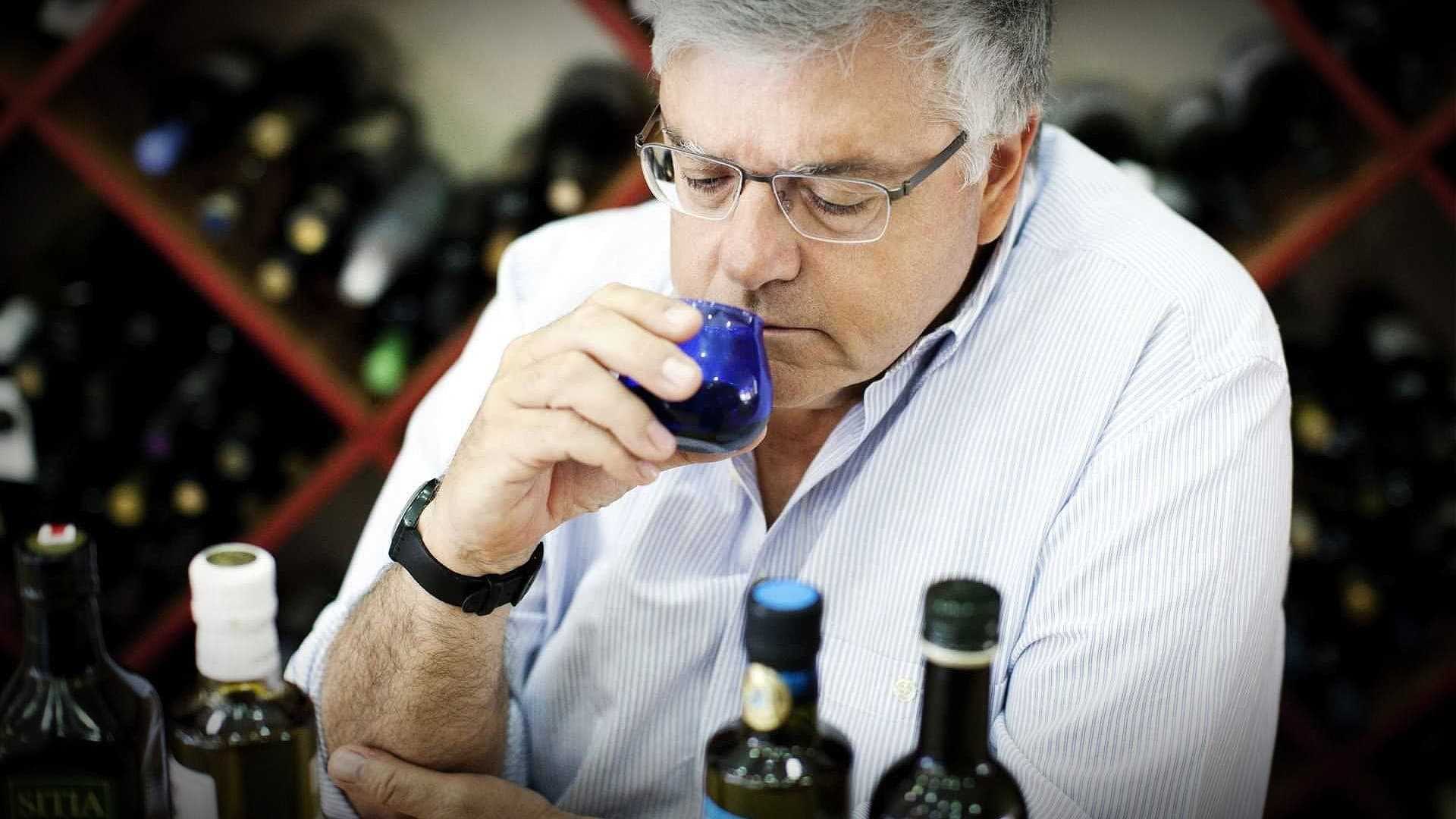 Ein Mann mit Brille riecht Olivenöl aus einer kleinen blauen Tasse, während er an einem Tisch mit Flaschen sitzt. - Olive Oil Times