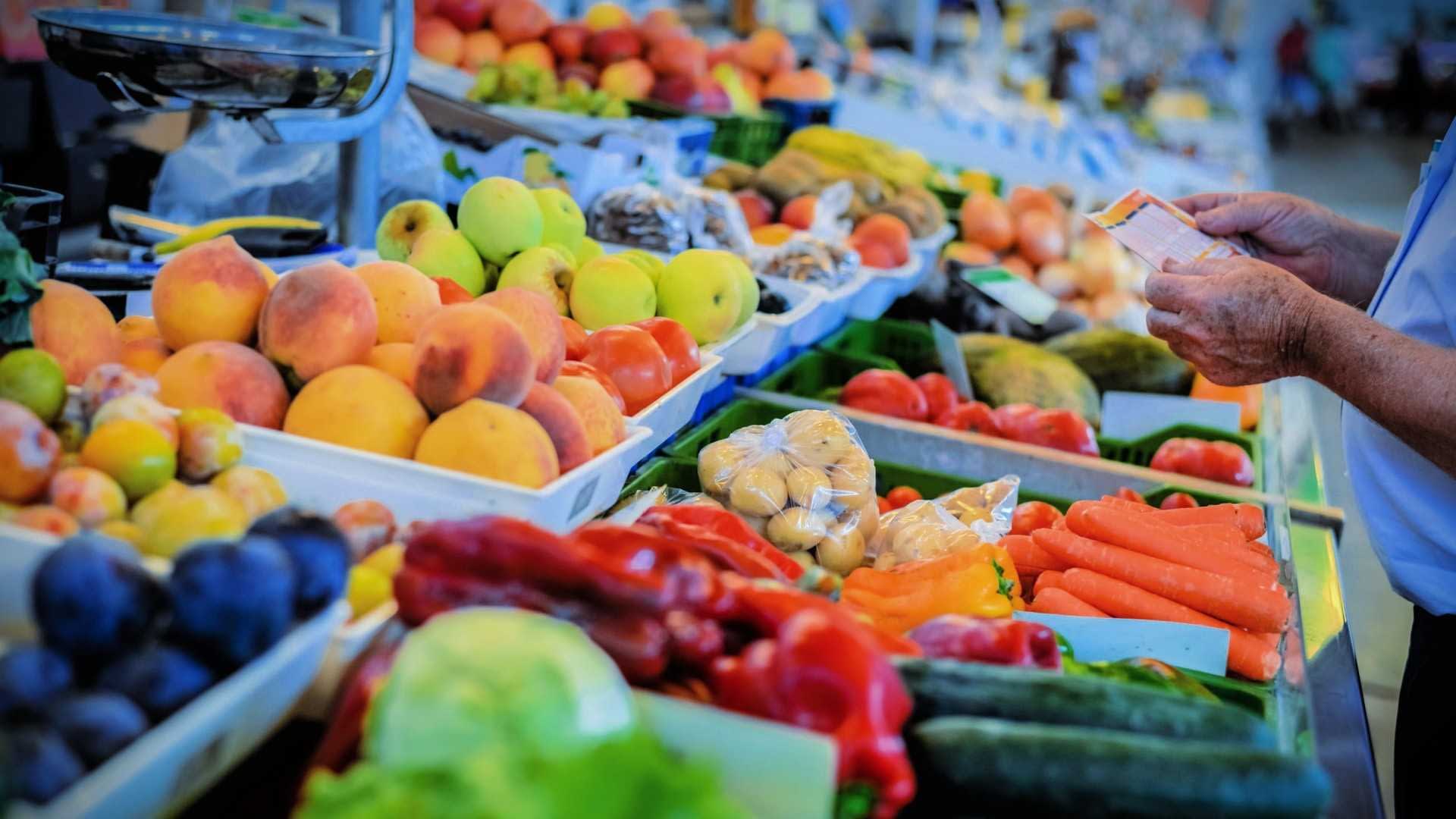 Eine Auswahl an frischem Obst und Gemüse, darunter Pfirsiche, Karotten und Gurken, wird an einem Marktstand präsentiert. - Olive Oil Times
