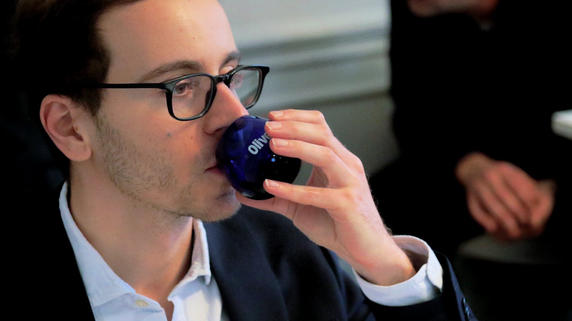 Un homme portant des lunettes buvant dans un verre bleu avec le mot « Olive » dessus. - Olive Oil Times
