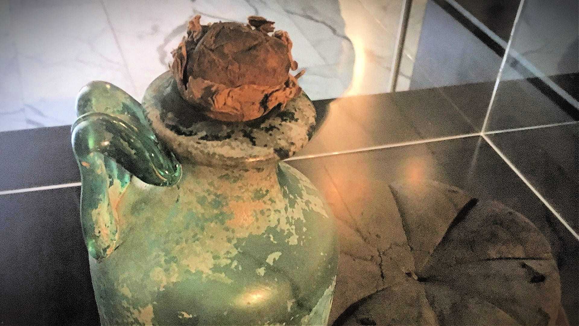 コルク栓が付いた古い緑色のガラスの水差しが表面に展示されています。 - Olive Oil Times