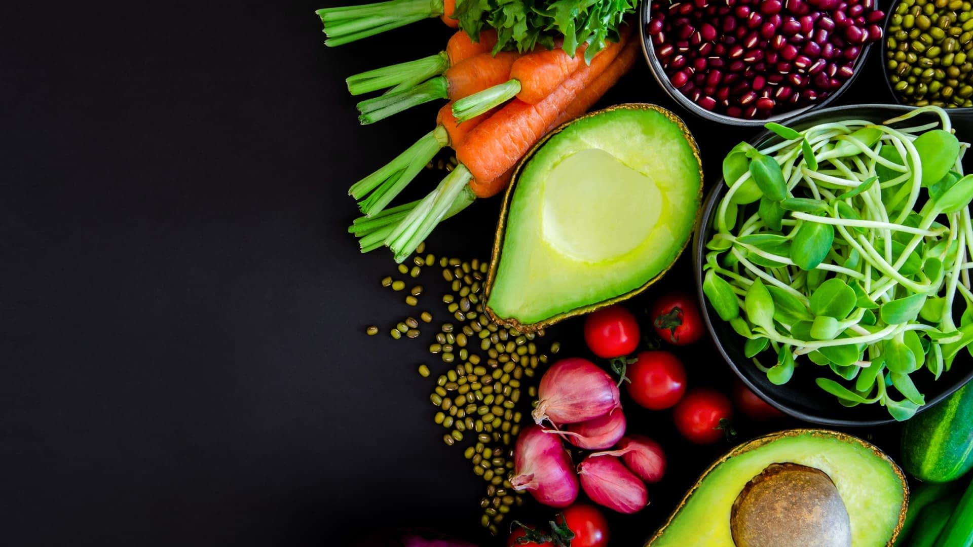 Uma variedade de vegetais frescos, incluindo cenouras, aipo e abacate, dispostos sobre uma superfície escura. Olive Oil Times