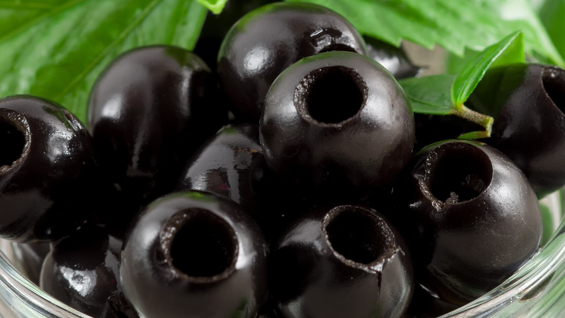 Primer plano de aceitunas negras con una superficie brillante, colocadas en un recipiente transparente con hojas verdes. - Olive Oil Times