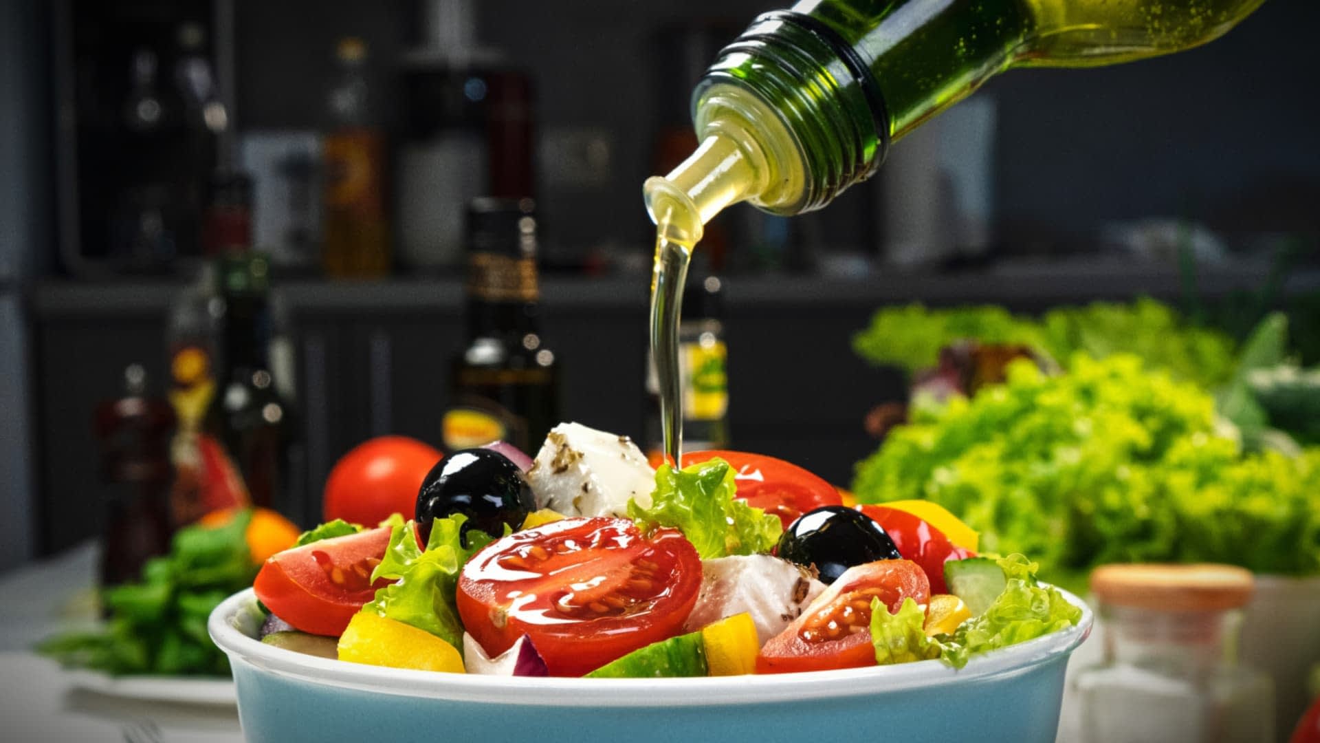 Aceite de oliva vertido sobre una ensalada fresca con tomates, lechuga y aceitunas en un bol. Olive Oil Times