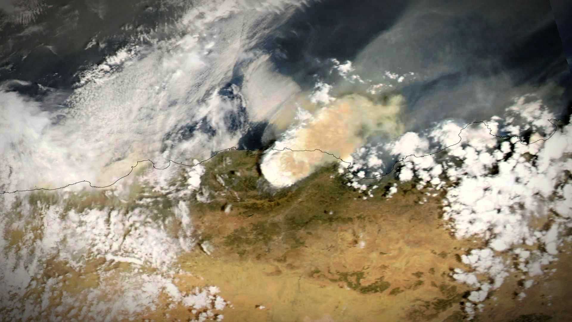 Спутниковый снимок, на котором виден дым, поднимающийся от лесного пожара, на фоне пейзажа с облаками и рельефом местности. Olive Oil Times