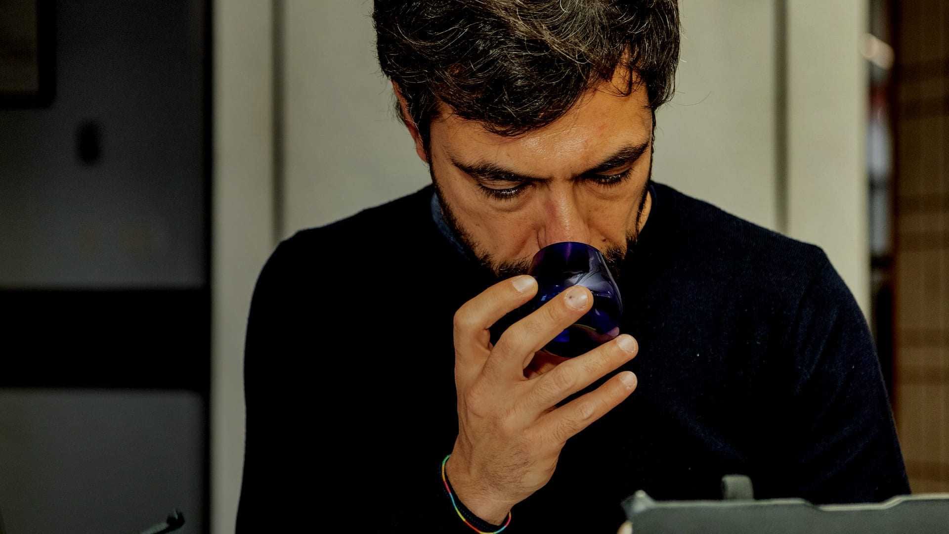 A man with dark hair and a beard holding a blue glass cup close to his face. - Olive Oil Times