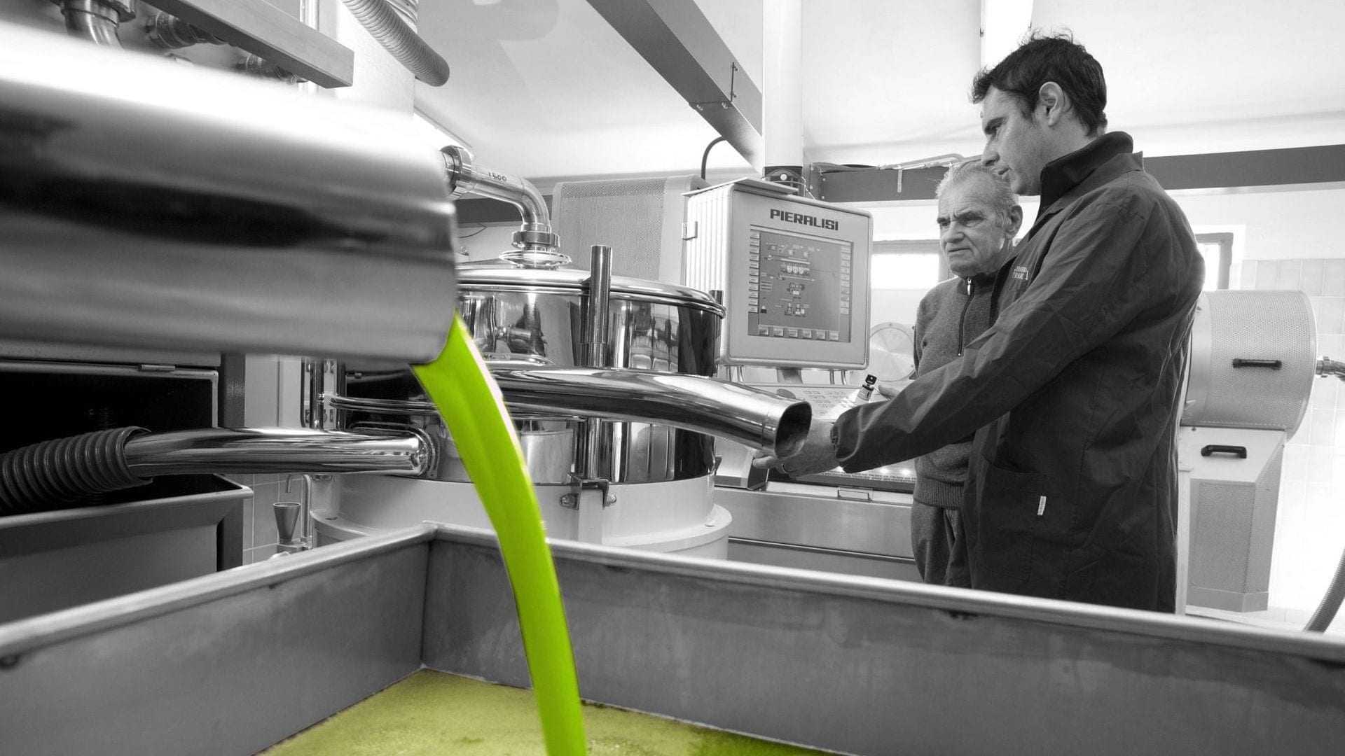 Two men observing the olive oil extraction process in a production facility with green liquid flowing. - Olive Oil Times