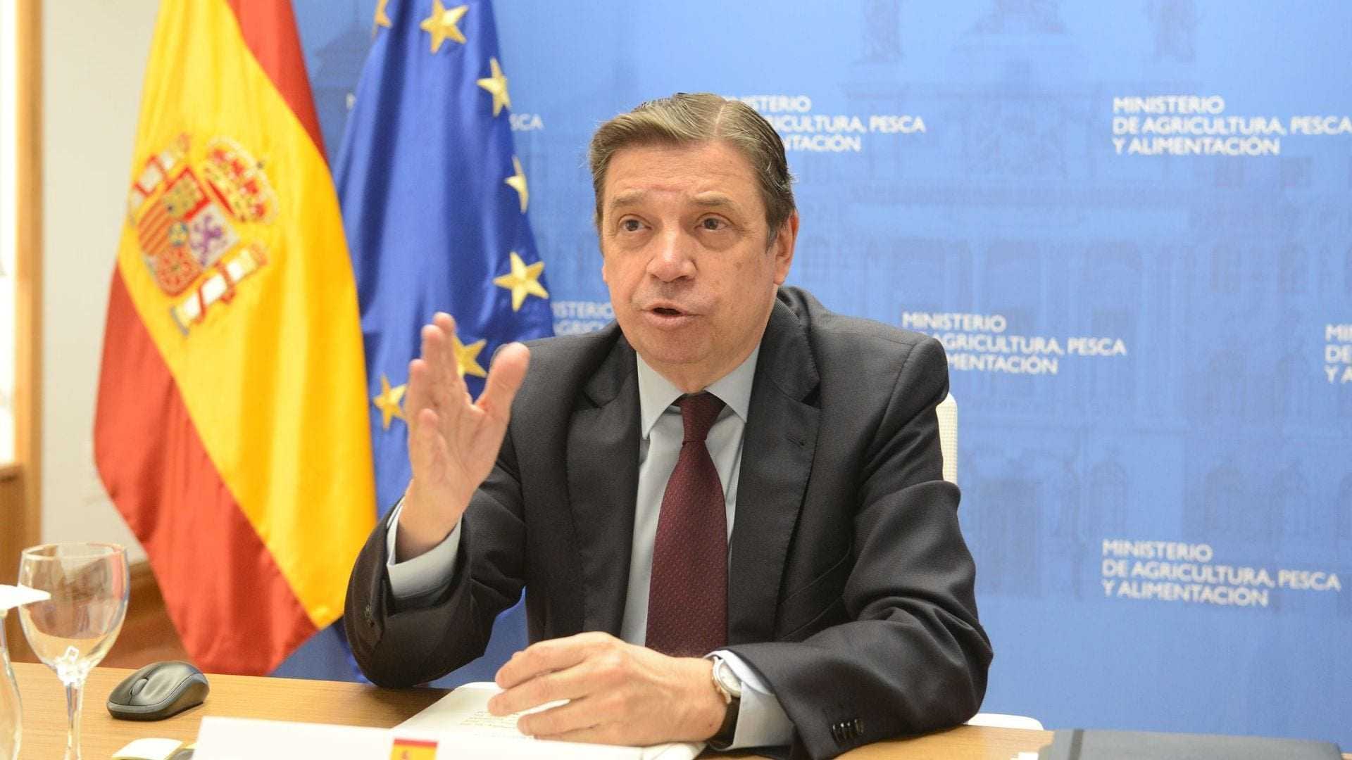 Luis Planas, a government official, speaking at a press conference with flags in the background. - Olive Oil Times
