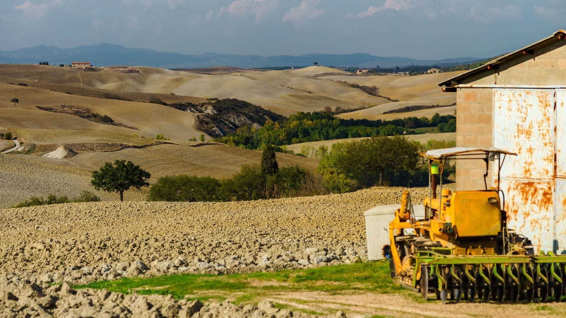 Yellow tractor parked near a barn in a rural agricultural landscape with rolling hills. - Olive Oil Times