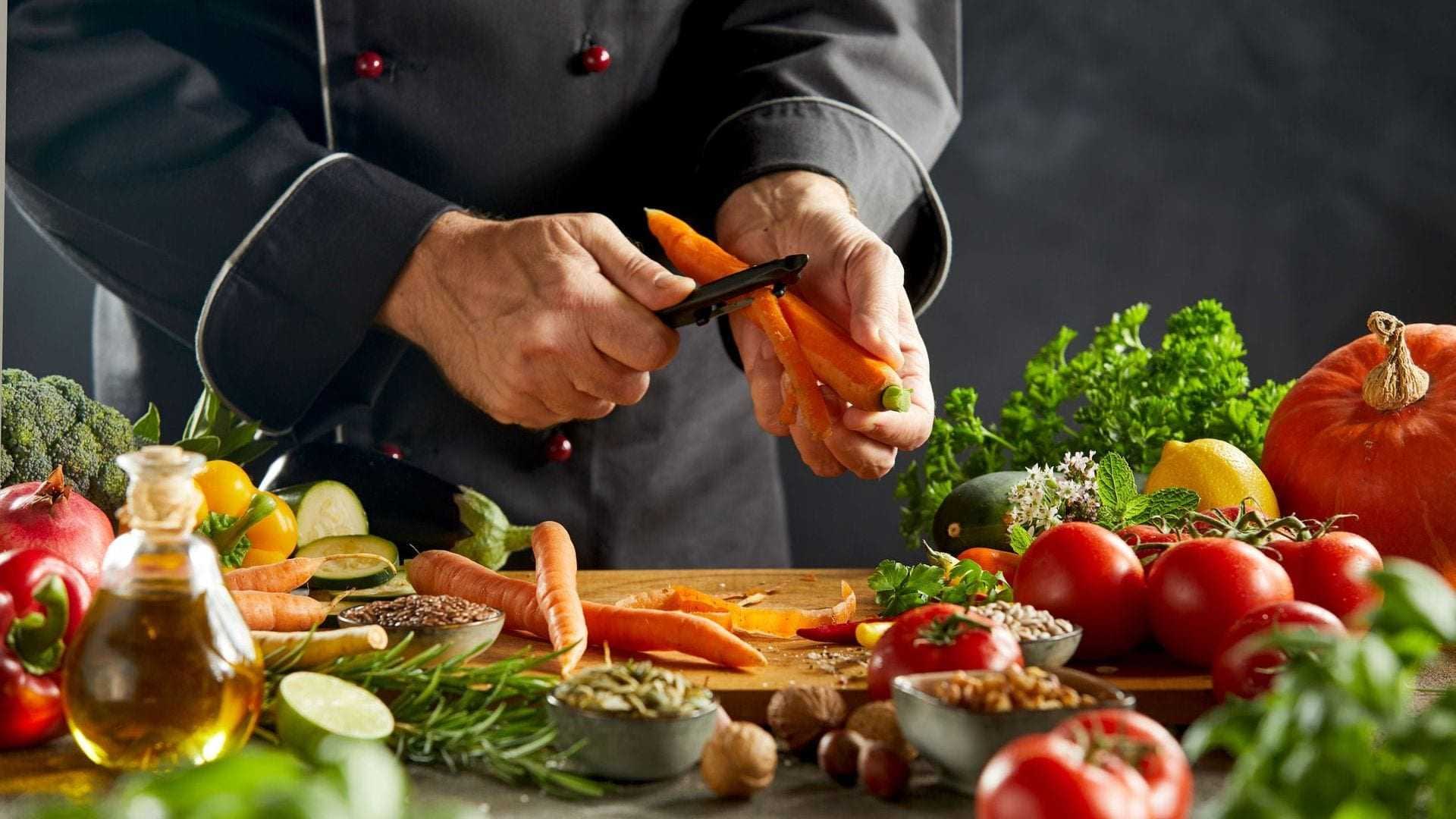 Chef peeling a carrot with various vegetables and herbs on a kitchen counter. - Olive Oil Times