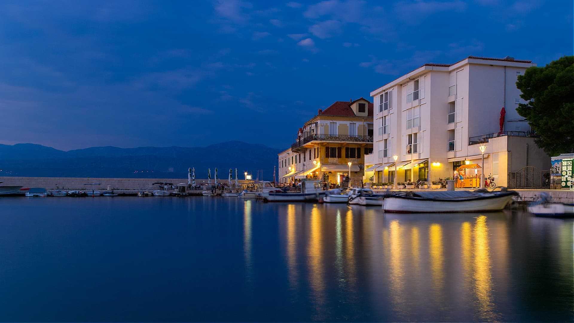 Waterfront scene featuring boats docked near a building with lights reflecting on the water at dusk. - Olive Oil Times