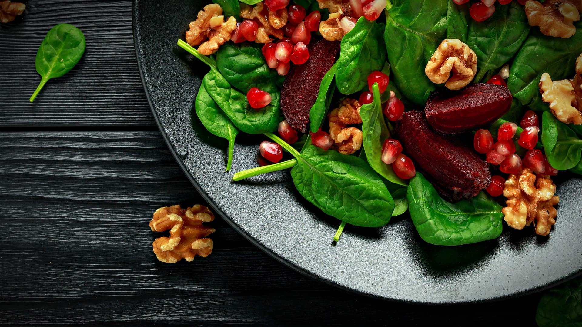 A black plate filled with spinach leaves, pomegranate seeds, and walnuts arranged together. - Olive Oil Times