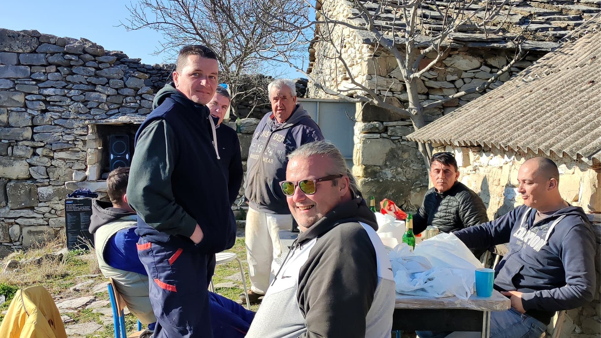 A group of six men gathered around a table outdoors, with stone buildings in the background. - Olive Oil Times