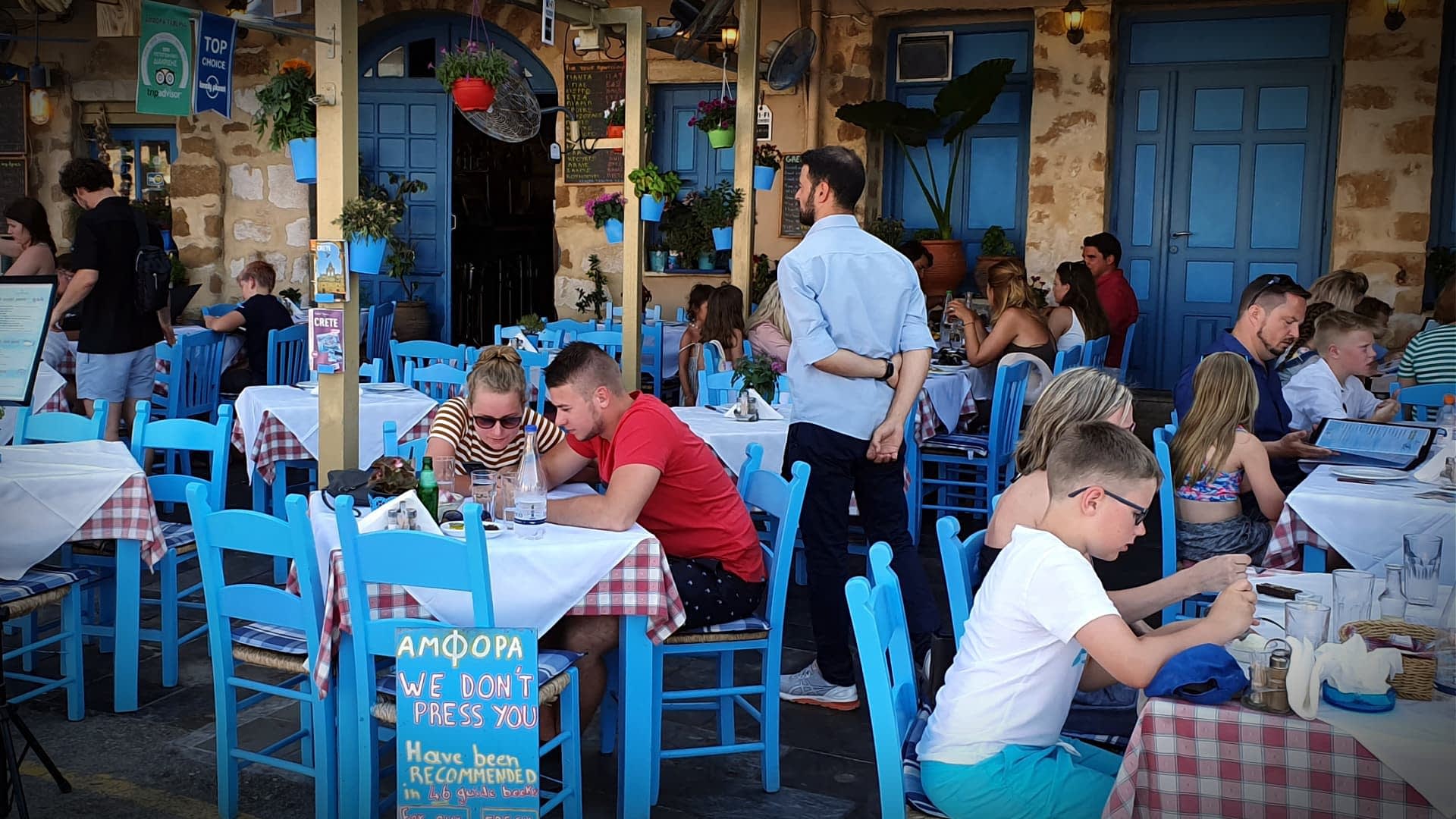 Outdoor dining area with tables and blue chairs at a restaurant, featuring patrons engaged in conversation. - Olive Oil Times