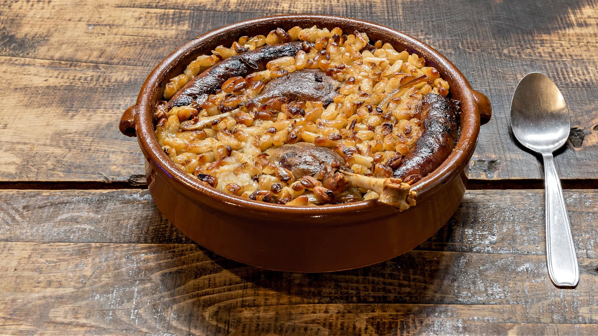 A baked dish containing sausages, onions, and grains in a round ceramic container on a wooden surface. - Olive Oil Times
