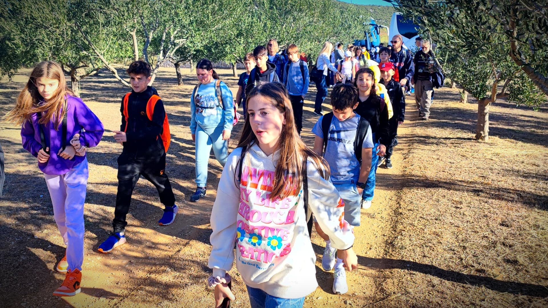 A group of students walking in an olive grove, wearing backpacks and casual clothing. - Olive Oil Times