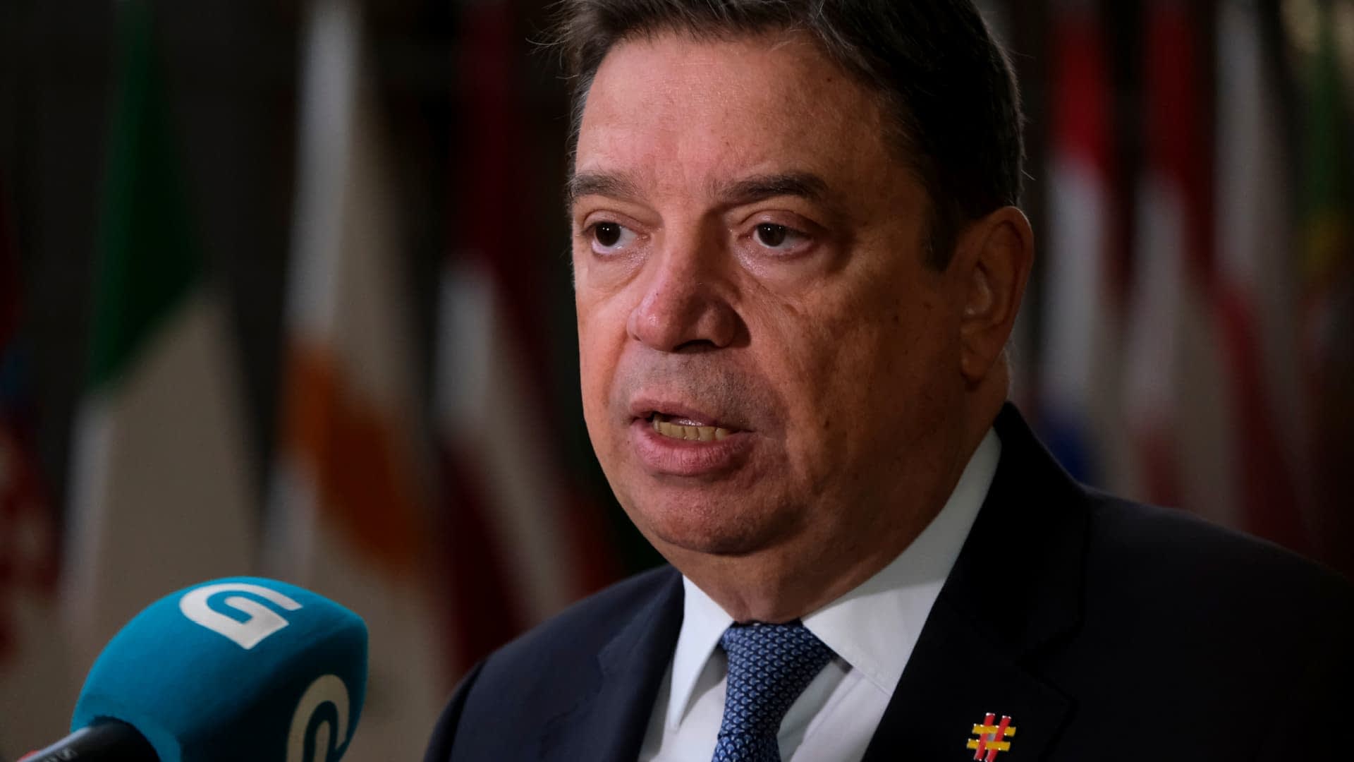 A man in a suit speaking at a press conference with flags in the background. - Olive Oil Times