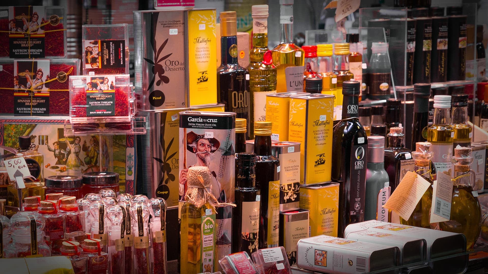 Various bottles and jars of olive oil and condiments displayed on a shelf in a market. - Olive Oil Times