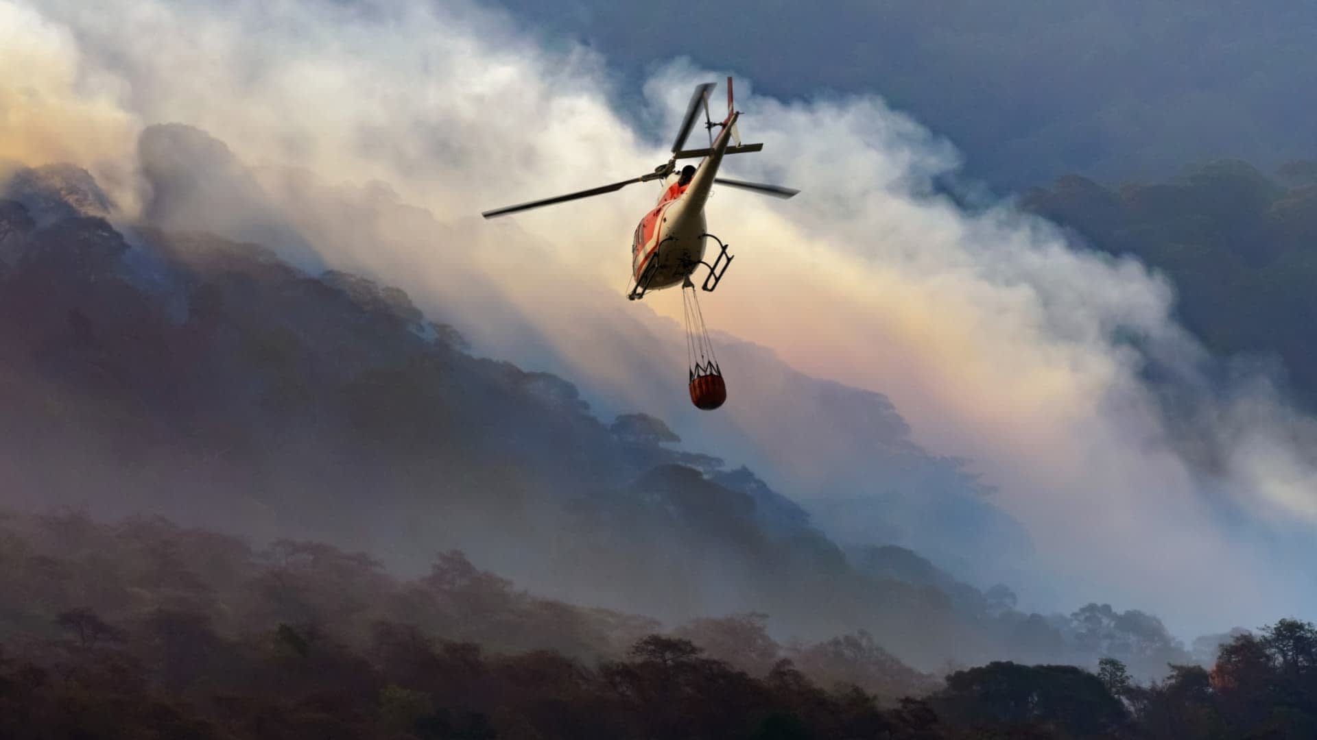 Helicopter flying over a mountainous area with a water bucket suspended below it. - Olive Oil Times