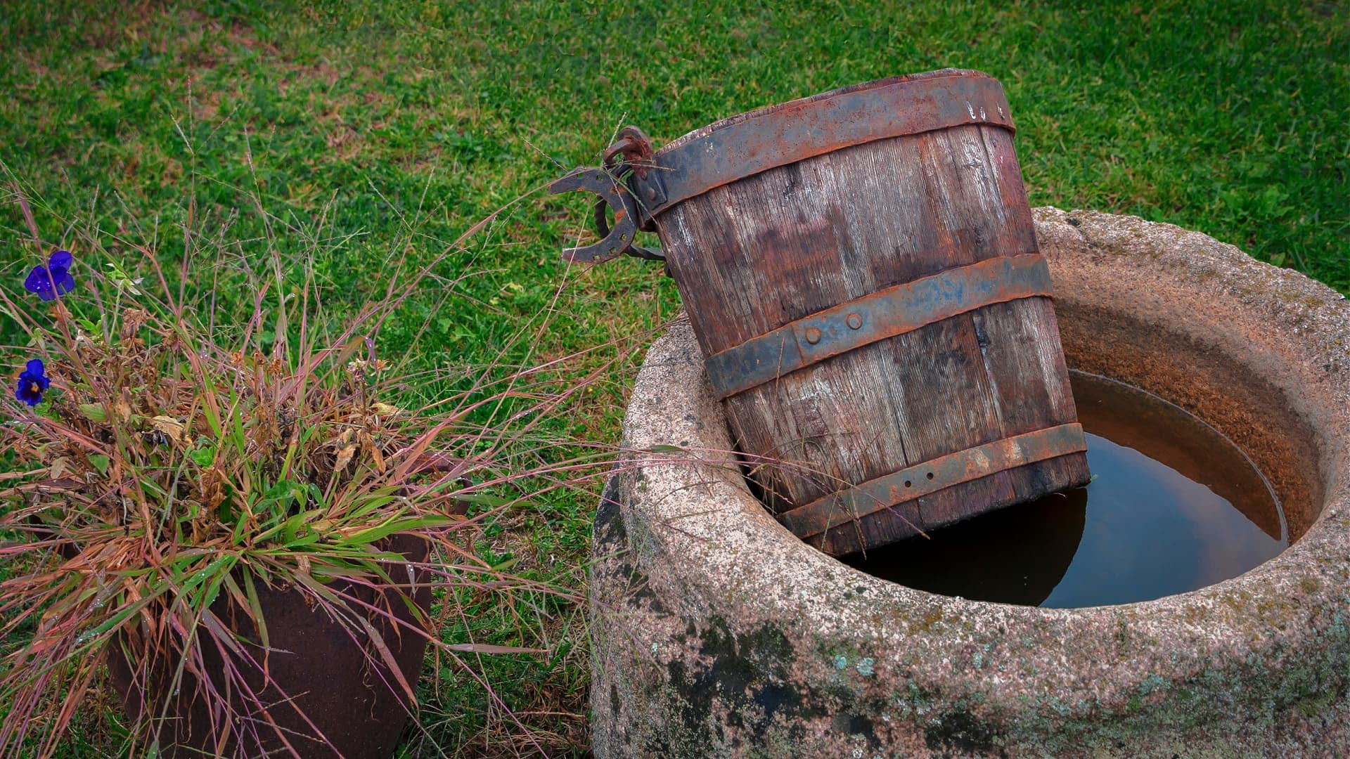 A wooden bucket partially submerged in a stone well filled with water. - Olive Oil Times