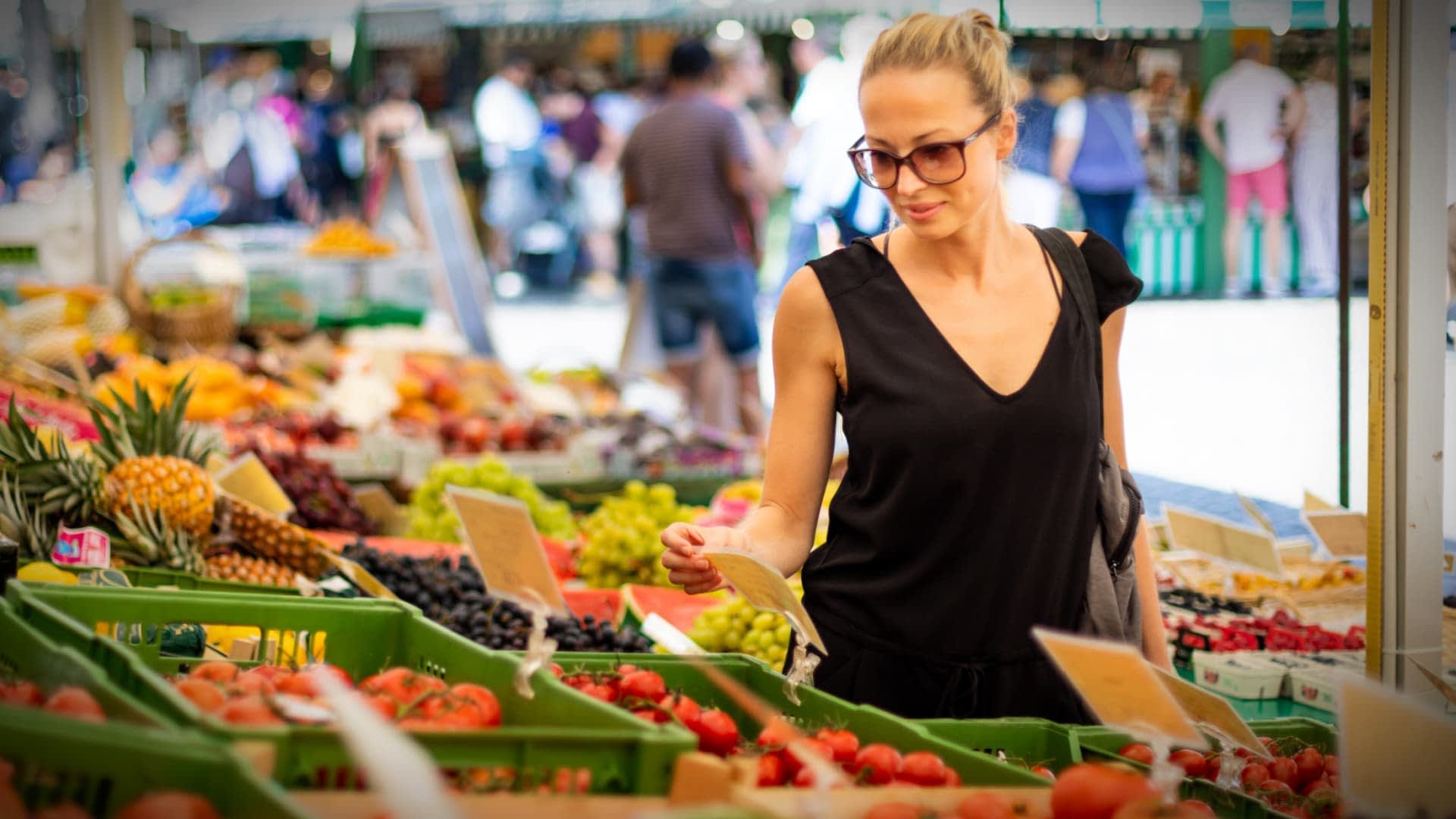 A woman wearing sunglasses examines produce at a farmers market with various fruits and vegetables displayed. - Olive Oil Times