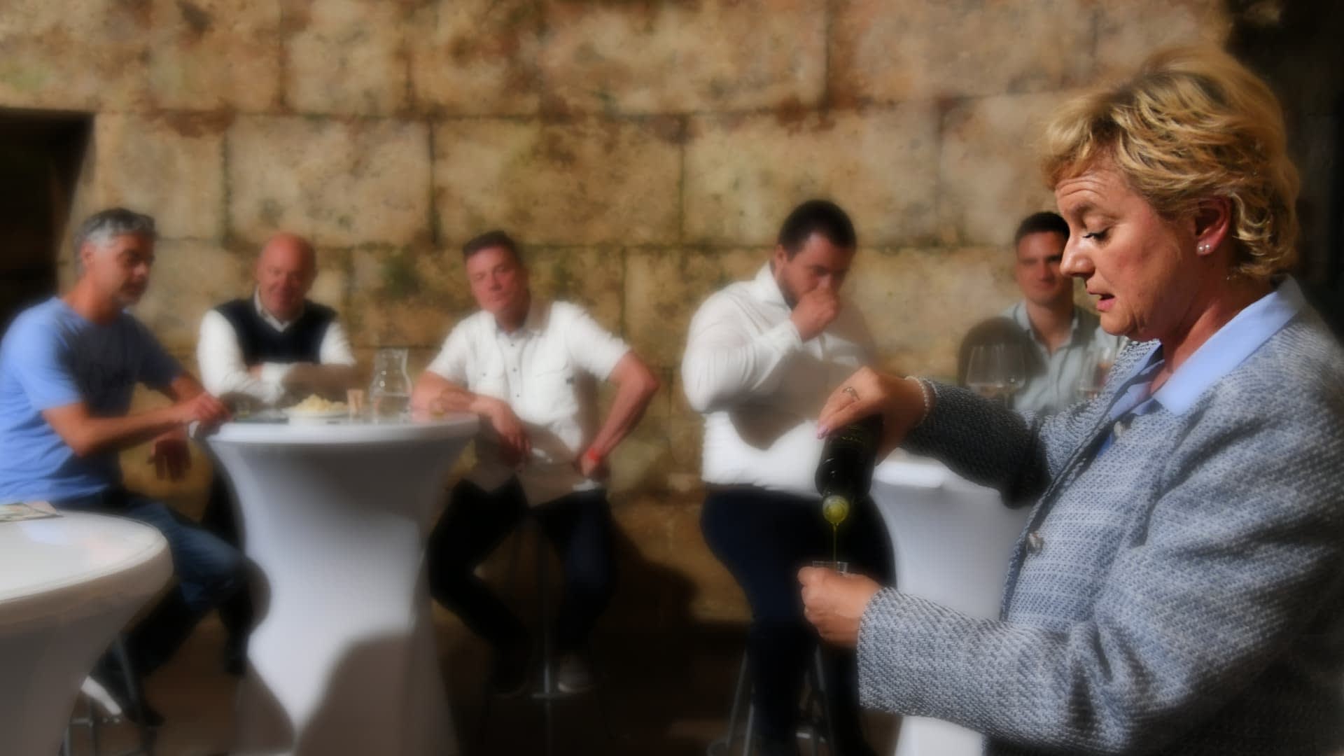 A woman in a gray blazer pouring olive oil while a group of men observes at a tasting event. - Olive Oil Times