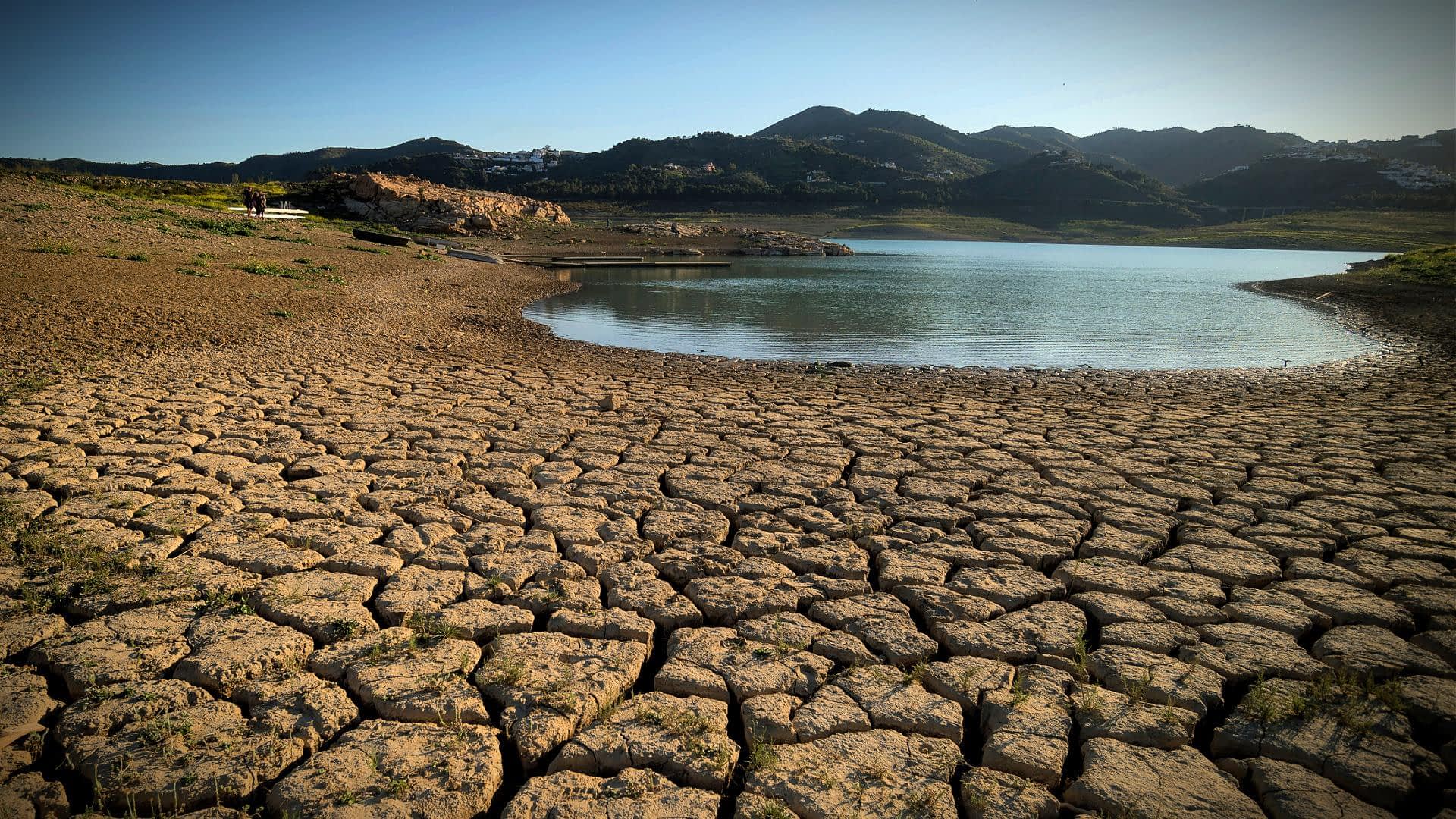 Dry, cracked earth surrounding a small body of water with mountains in the background. - Olive Oil Times