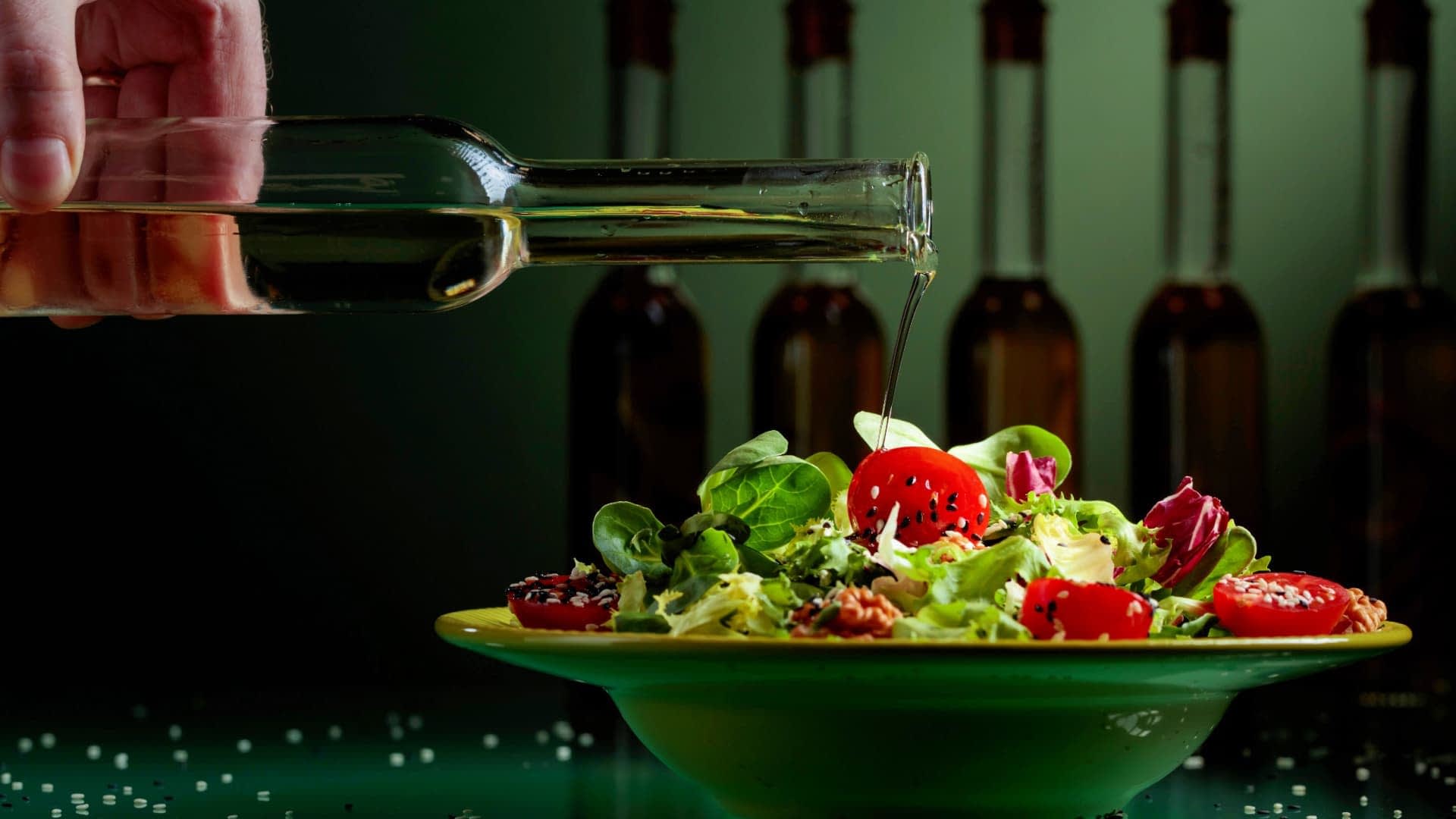 A hand pouring olive oil from a glass bottle onto a salad in a green bowl with cherry tomatoes and greens. - Olive Oil Times